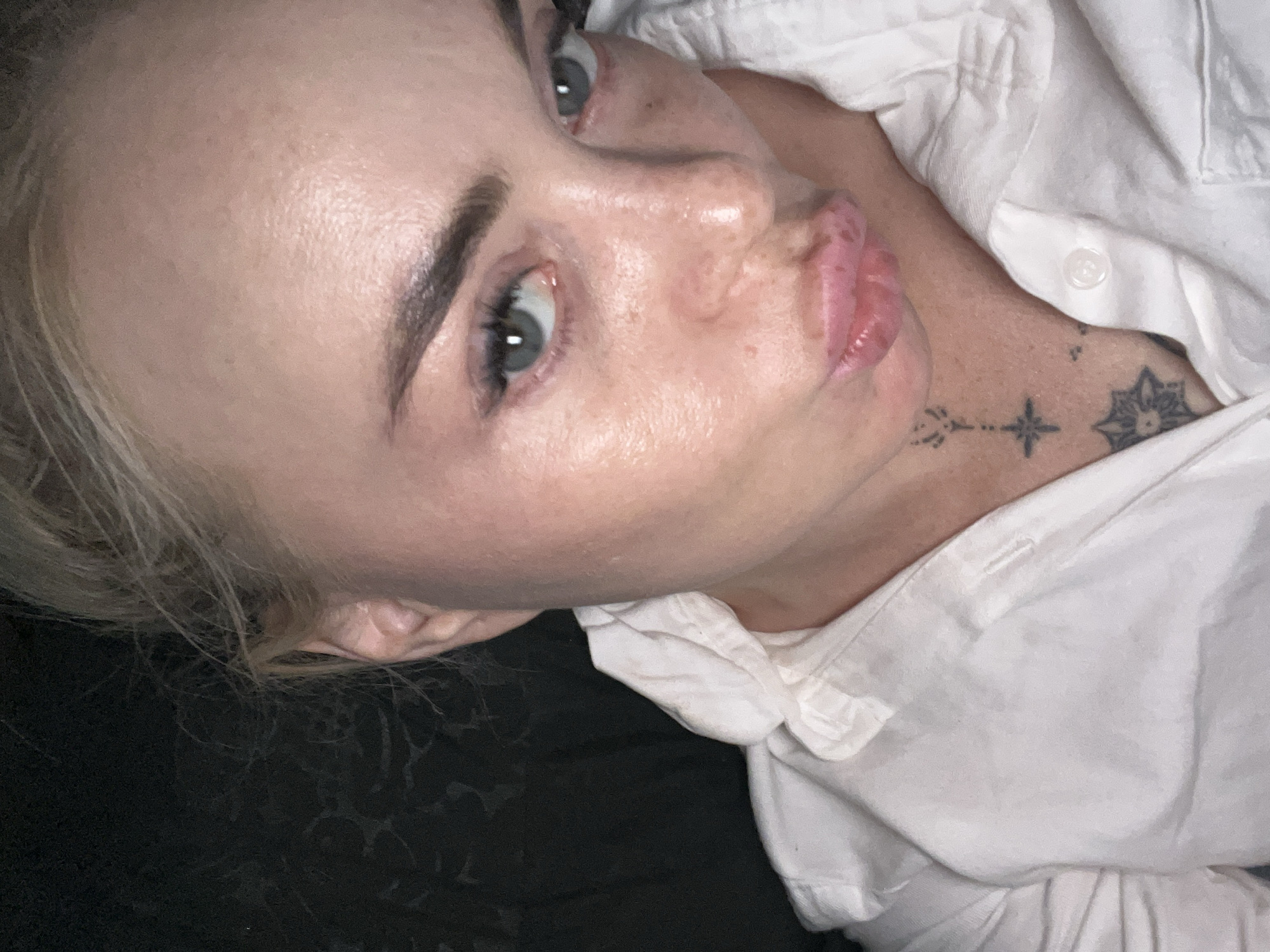 A medium shot shows a close-up of a woman's face, angled slightly to the right, with her lips puckered. She has blonde hair pulled back, visible at the top of the frame, and is wearing a white collared shirt. The background is dark and appears to be a textured surface. Tattoos are visible on her neck. The lighting is dim, casting shadows.