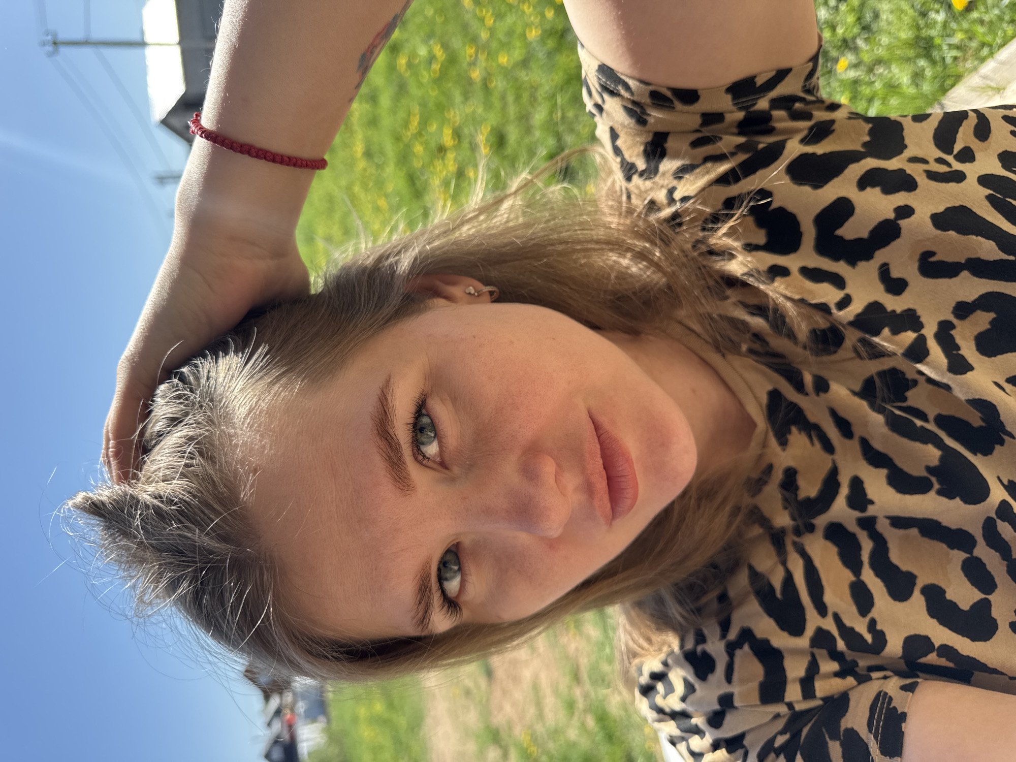 A woman is outdoors in the city of Ферма, Russia. She is fair-skinned with light brown hair and light eyes. She is wearing a tan and black leopard print top. A red braided bracelet is on her left wrist. Her left hand is on her head. There is grass and yellow flowers visible in the background. A blue sky is also in the background. A building and car can be seen blurred in the distance.