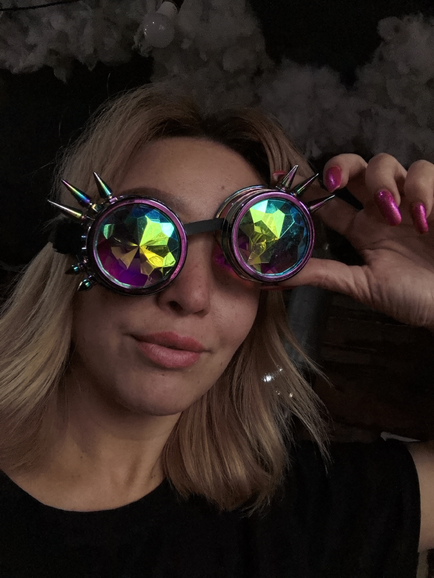 A fair-skinned woman with blonde hair is holding up a pair of steampunk goggles with faceted lenses and spiked edges, the goggles are pink and black. She has pink nails and is wearing a black shirt. The background is dark, with a light bulb and what appears to be artificial fog. There is no information to determine if the location is Noviy, Russia.