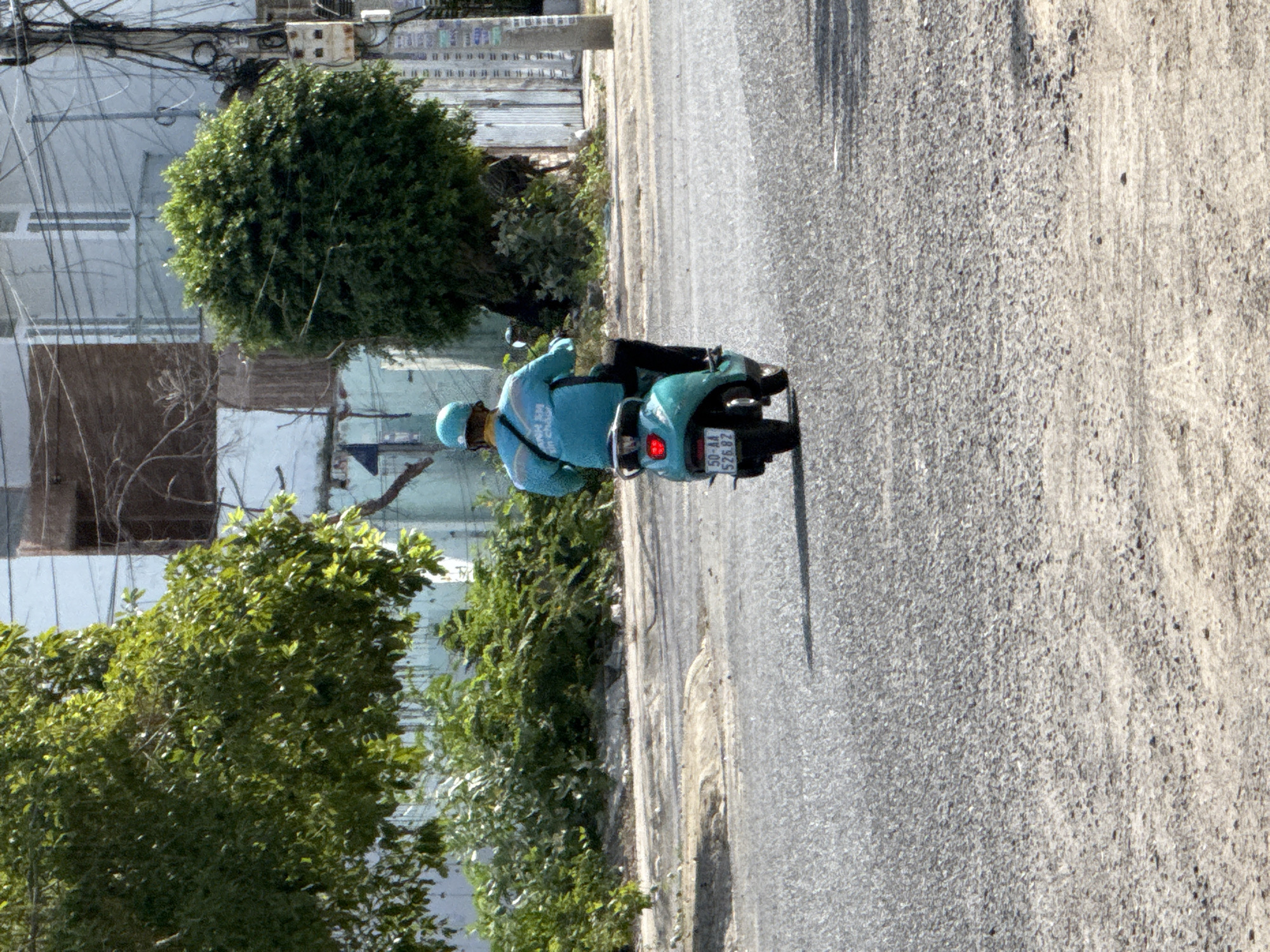 In the city of Bac Nha Trang Ward, Vietnam, a person wearing a turquoise helmet and a matching turquoise long-sleeved shirt with black pants rides a turquoise scooter on a paved road. The scooter has a license plate that reads "50-AA 526.82". Behind the rider, the scooter's brake light is illuminated red. The road appears to be in good condition with some texture and debris. To the left, there is a large green tree with lush foliage, and to the right, another dense green tree stands. In the background, a multi-story building with white and blue walls is visible, along with a utility pole and a network of electrical wires. The scene is bathed in bright sunlight, casting shadows on the road.