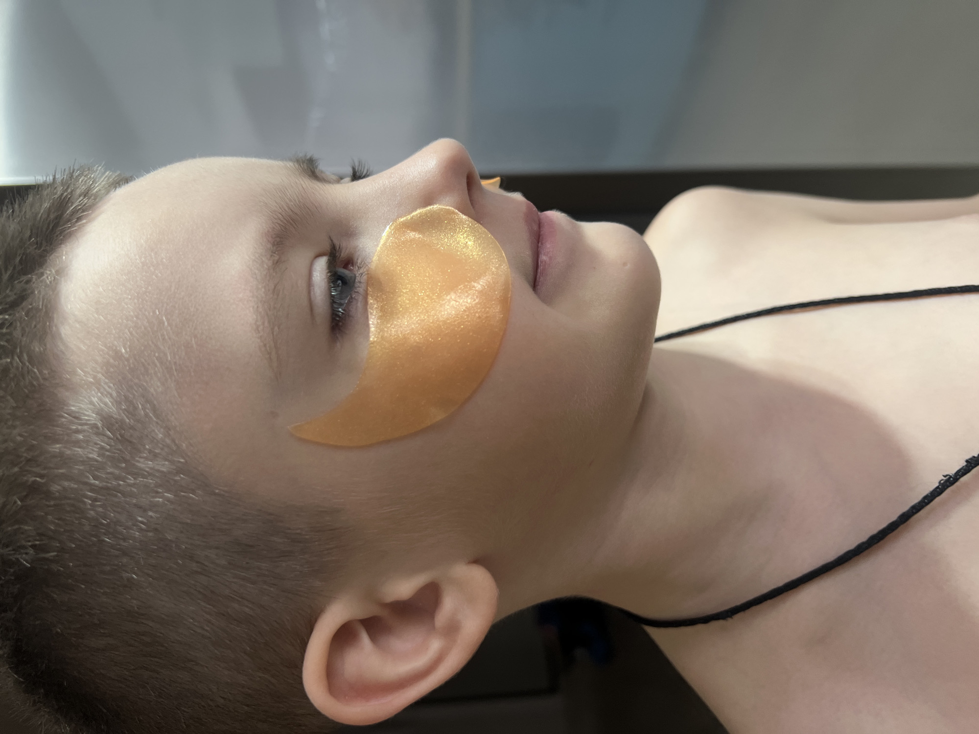 A young person lies on a dark surface, eyes closed, with a golden eye patch placed under one eye. A thin, dark necklace rests on their chest. The location is suspected to be Марковский, Russia.