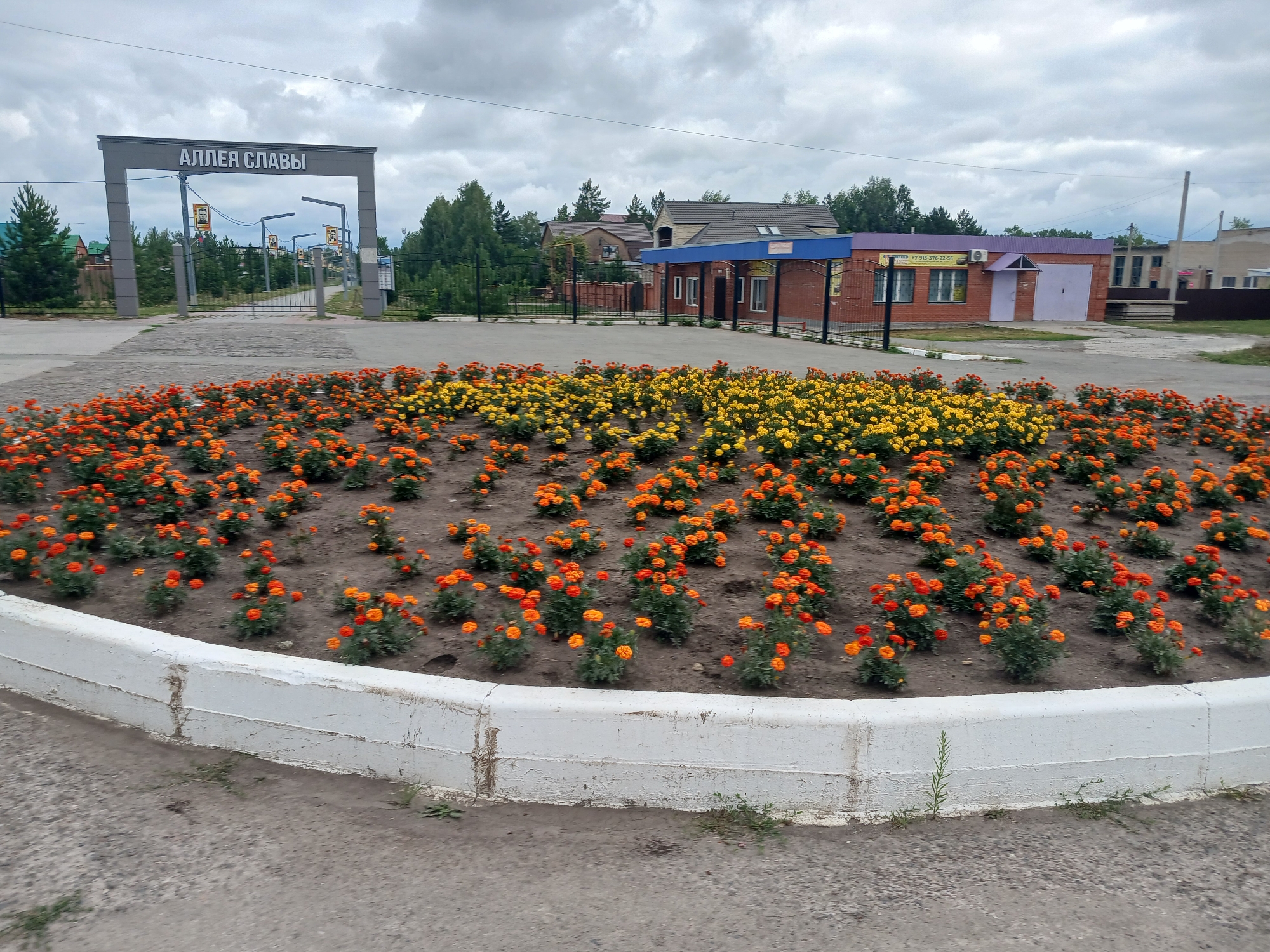 In Prokudskoye, Russia, a bright flowerbed featuring a profusion of orange and yellow marigolds greets the viewer in the foreground. The flowers are planted in neat rows, creating a vibrant splash of color against the muted tones of the surrounding environment. Behind the flowerbed, an archway stands proudly, bearing the inscription "АЛЛЕЯ СЛАВЫ" (Alley of Glory) in bold white lettering. Beyond the arch, a paved pathway leads into the distance, flanked by trees and fencing. Several buildings, likely commercial or residential, are visible in the background, their brick facades and pitched roofs contributing to the semi-urban landscape. The sky above is overcast, with heavy clouds obscuring the sun and casting a soft, diffused light on the scene. The overall impression is one of a well-maintained public space, likely a park or memorial area, designed to be both beautiful and commemorative.