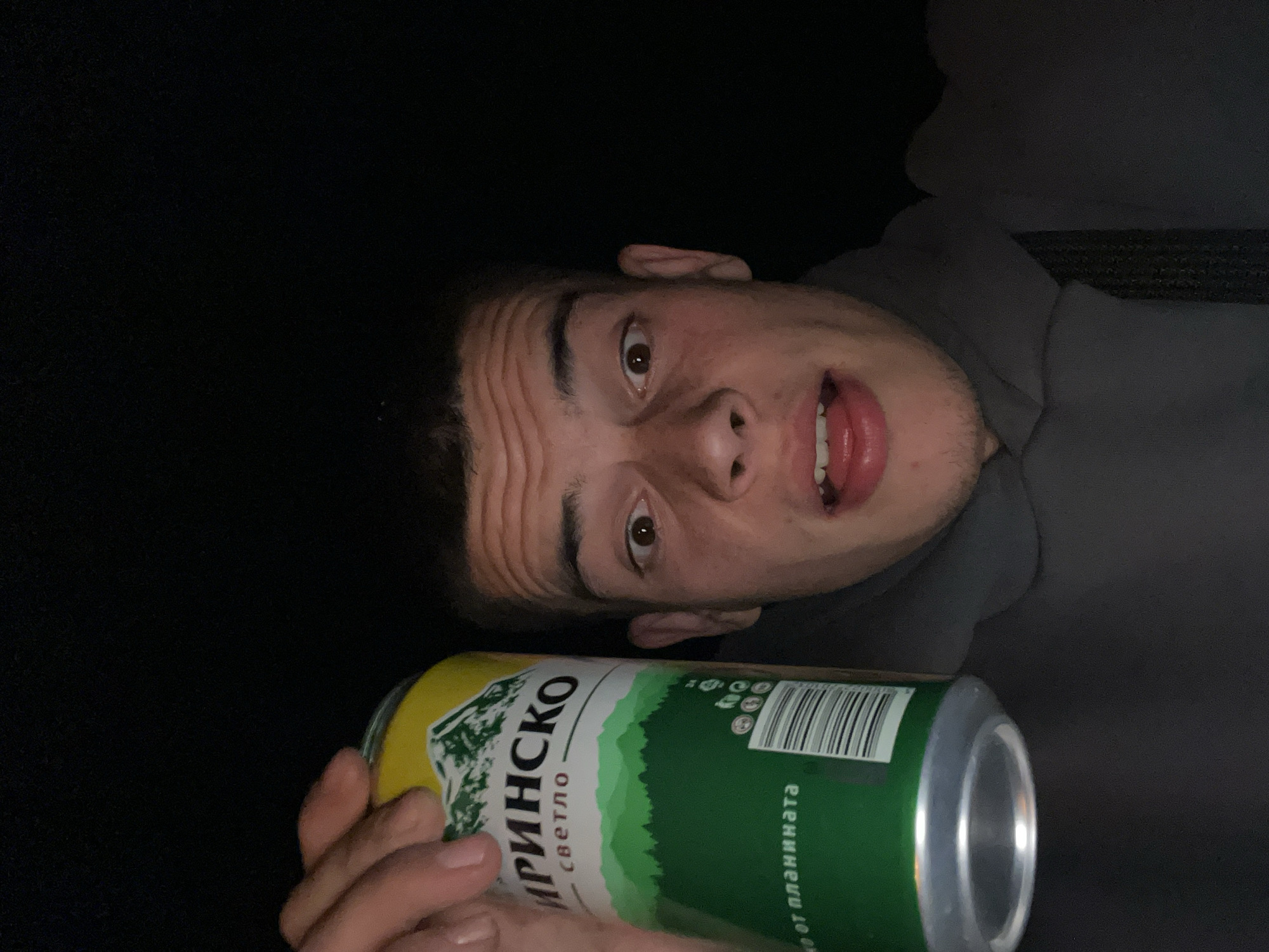 A person is holding a can of "PIRINSKO" beer, which is a Bulgarian beer brand. The can has a green and white design featuring a mountain landscape. The background is dark, and the person is wearing a grey hoodie. The image is taken from a low angle. The city of Karavelovo, Bulgaria is not apparent from the image.