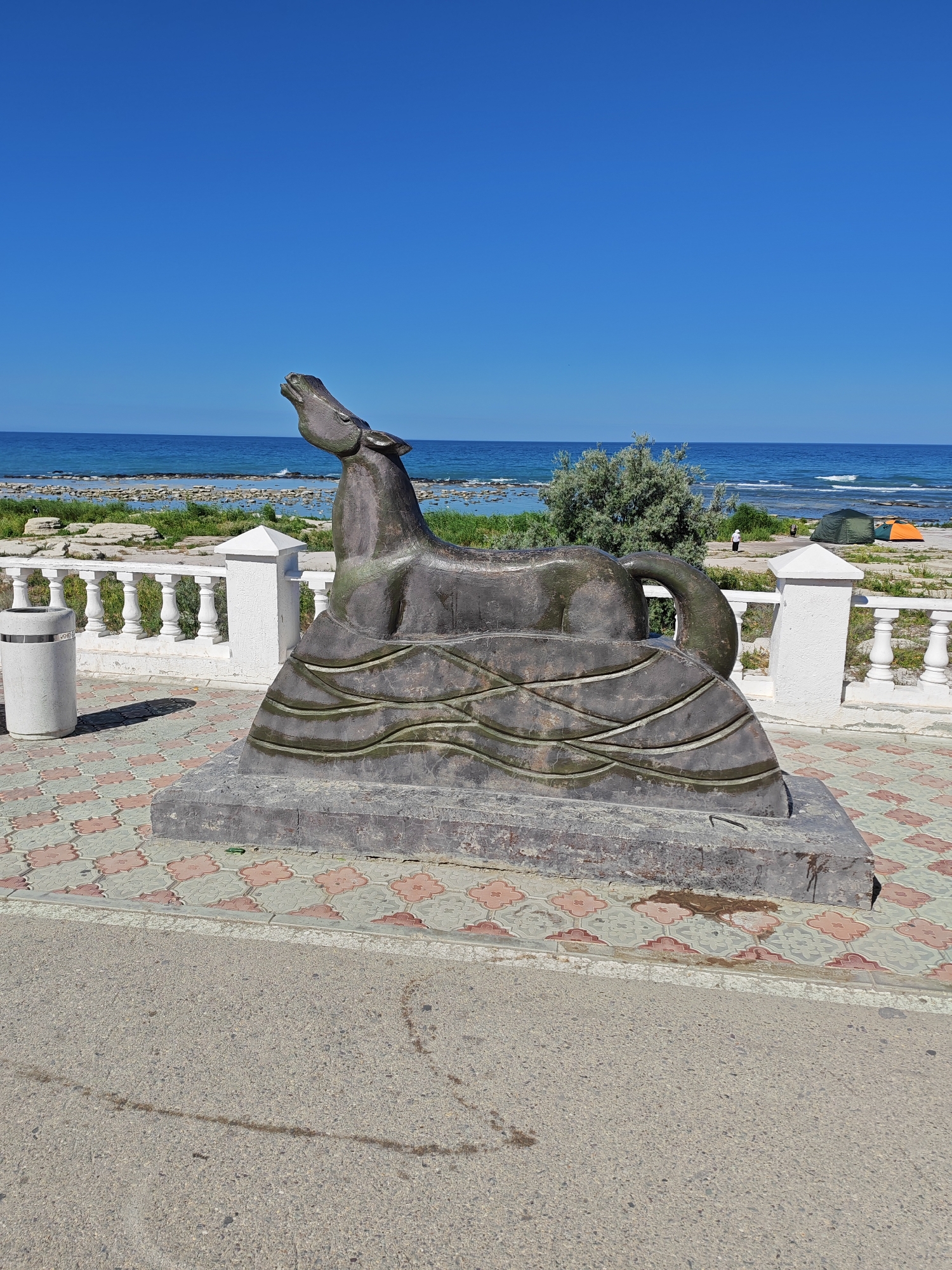 The image depicts a bronze sculpture of a horse emerging from stylized waves, situated on a stone pedestal. The sculpture is located outdoors, along a promenade next to the Caspian Sea. In the background, the sea is visible under a clear blue sky, with a rocky shoreline and sparse vegetation. A white railing runs along the promenade, separating it from the beach area. To the left of the sculpture, a grey trash receptacle is positioned. The ground in front of the sculpture is paved with decorative tiles in a repeating pattern of pink and beige floral shapes, bordered by asphalt. The scene appears to be a public space in Aktau, Kazakhstan, likely a tourist area by the sea.
