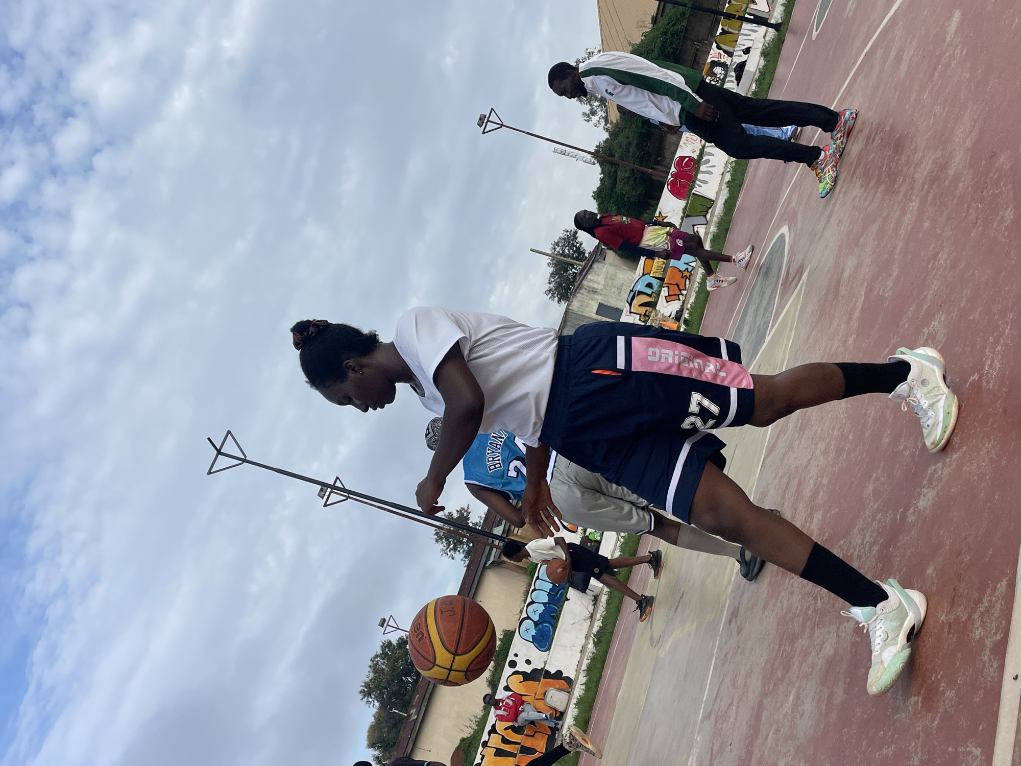 The image depicts an outdoor basketball court, likely located in Jos, Nigeria, under a cloudy sky. In the foreground, a young woman in a white t-shirt and navy blue shorts with pink trim, identified by the number "27" on her shorts, is in motion, her right arm raised as if to pass or shoot a basketball. A basketball is suspended in the air to her left.
Several other individuals are present on the court. To the left of the foreground woman, another player in a blue jersey with the number "24" and "BRYAN" is visible. Further in the background, to the right, a man in a white and green jacket and black pants is standing. On the far left, a person in a red jacket is seated on a bench adorned with graffiti.
The court surface is red and marked with white lines. Behind the court, a wall covered in vibrant graffiti is visible, with buildings and trees in the distance. Basketball hoops and poles are also present, indicating the recreational use of the area. The overall scene captures a moment of athletic activity and casual social interaction within an urban setting.