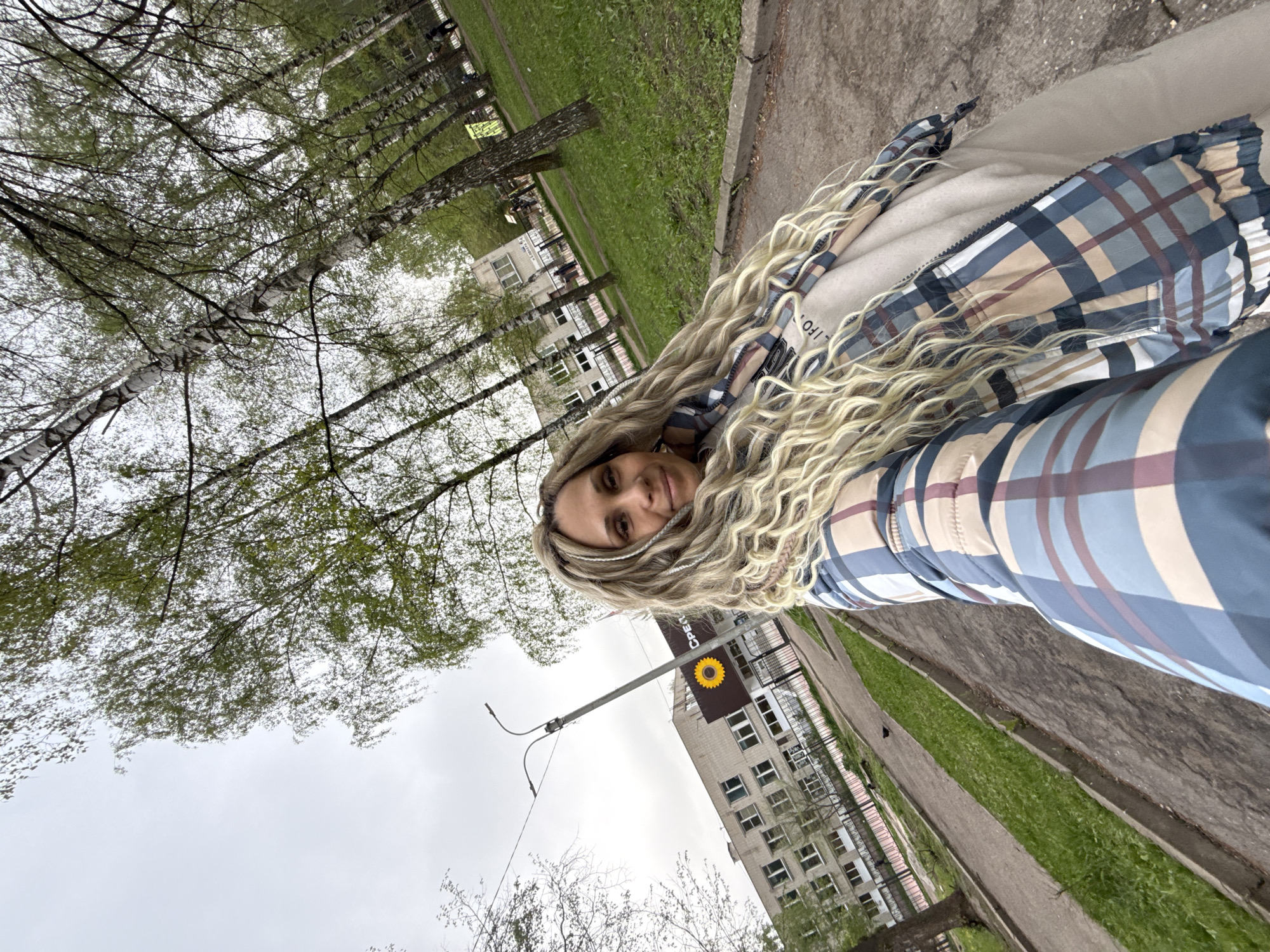 A woman is photographed at an acute angle from below, facing the camera, with her head tilted slightly towards her right. Her blond hair, styled in curls, cascades around her head and shoulders. She is wearing a plaid jacket with shades of blue, tan, and brown. The background indicates an outdoor setting. Above and to the left of the woman, there is a sparse canopy of trees, their slender trunks visible. Behind her, there is a light-colored multi-story building with a distinctive yellow sunflower emblem on its side. A street lamp is partially visible to the left of the building. The ground consists of a paved sidewalk, which transitions to a strip of grass further away. The location appears to be a city environment, likely within the city of Unknown, Unknown.