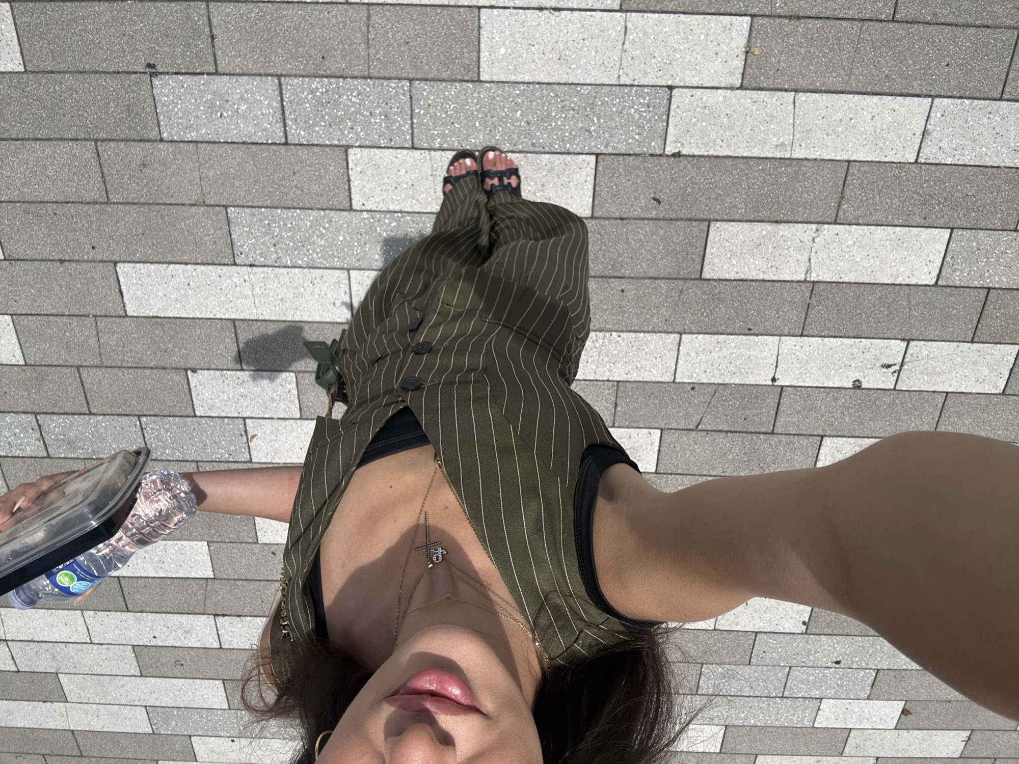 An overhead, high-angle full shot captures a person from the chest down, holding a clear plastic water bottle and a clear plastic container. The person is wearing a green pinstripe jumpsuit, a black bra, and black sandals. Their left arm is extended towards the camera. The ground is paved with rectangular grey and white tiles. The image is cropped to focus on the person and the immediate surroundings. The scene appears to be outdoors in an urban setting, likely a public walkway.