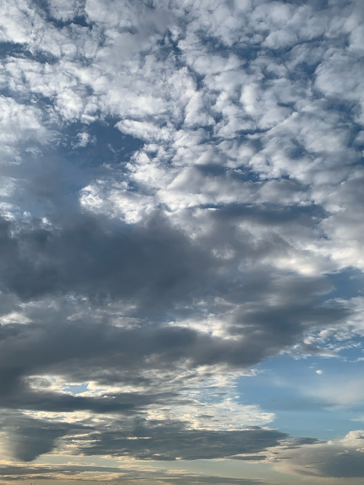 The image captures a dynamic sky over the city of Kolomyia, Ukraine. A complex layering of clouds dominates the scene. In the upper portion, scattered white cumulus clouds are interspersed with patches of blue sky. Mid-level, darker, more ominous clouds move across, creating shadows and texture. Towards the lower portion of the image, the clouds are denser and appear lower in the atmosphere, with some wispy formations and hints of sunlight breaking through. The horizon is faintly visible at the very bottom, suggesting land or buildings but lacking distinct detail. No people, subjects, or specific objects are identifiable within the frame. The overall impression is of an overcast day with varied cloud formations and changing light conditions.