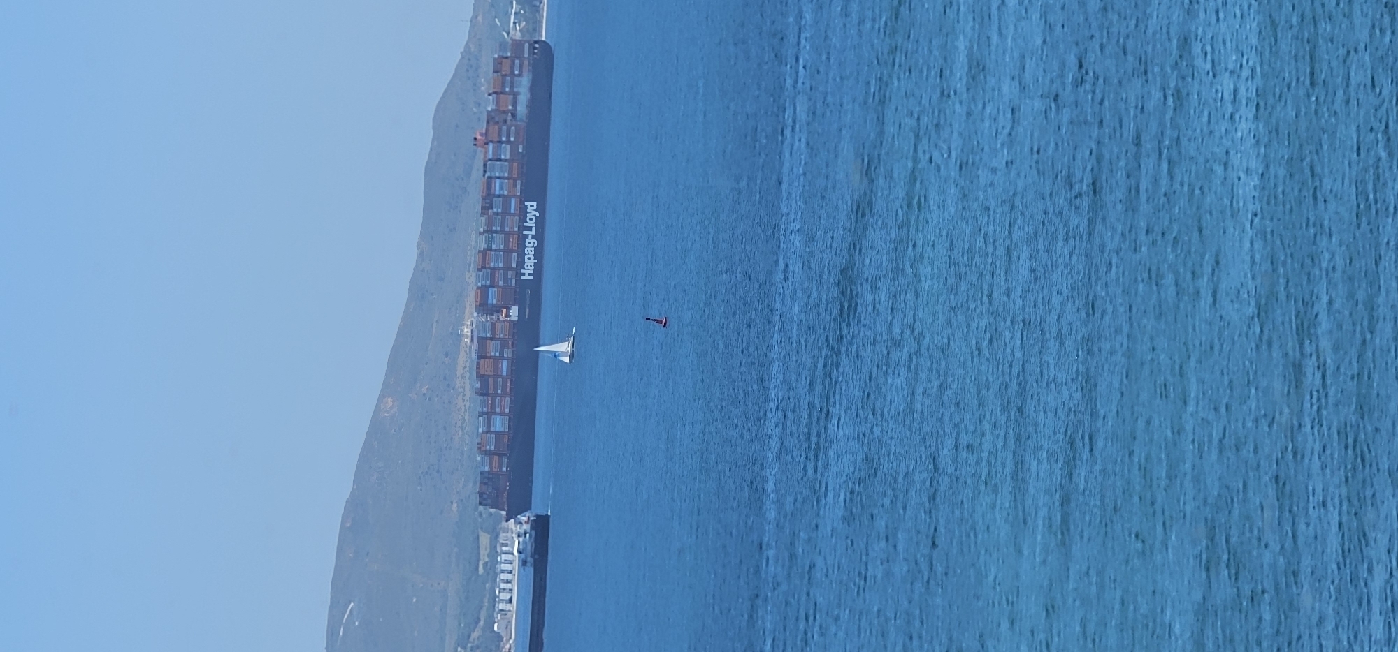 This image depicts a sunny daytime maritime scene near a mountainous coastline, likely in Algeciras, Spain. The dominant feature is a massive container ship, its side clearly displaying the text "Hapag-Lloyd." The ship is loaded with numerous multi-colored containers, indicating its role in global shipping, and appears to be navigating or docked along the base of a significant landmass.
To the right of the large vessel, a small, white-sailed sailboat gracefully cuts across the calm, blue water. Further out in the water, a distinctive red, cone-shaped navigational buoy marks a point on the surface. The water itself is a deep, clear blue, reflecting the bright conditions.
In the background, a large, rugged mountain or hill rises, providing a natural backdrop to the busy waterway. The sky above is a uniform, clear light blue, suggesting excellent weather conditions with no clouds. The overall impression is one of contrasting scales, with the enormous cargo ship sharing the waters with the much smaller sailboat, all under a clear sky in a scenic coastal environment.