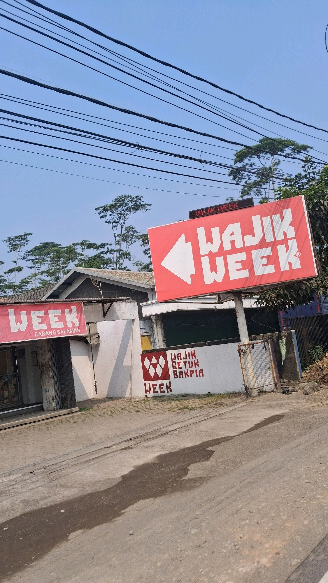 The image shows a street scene in Pabelan, Indonesia, with a focus on a red billboard advertising "WAJIK WEEK" with an arrow pointing left. Below the billboard, a white wall displays "WAJIK GETUK BAKPIA" and "WEEK" with a geometric logo. To the left, a smaller red banner reads "WEEK CABANG SALAMAN." The environment includes a road, power lines overhead, trees, and buildings. The lighting suggests it is daytime, possibly mid-morning or afternoon. There are no people visible in the frame.