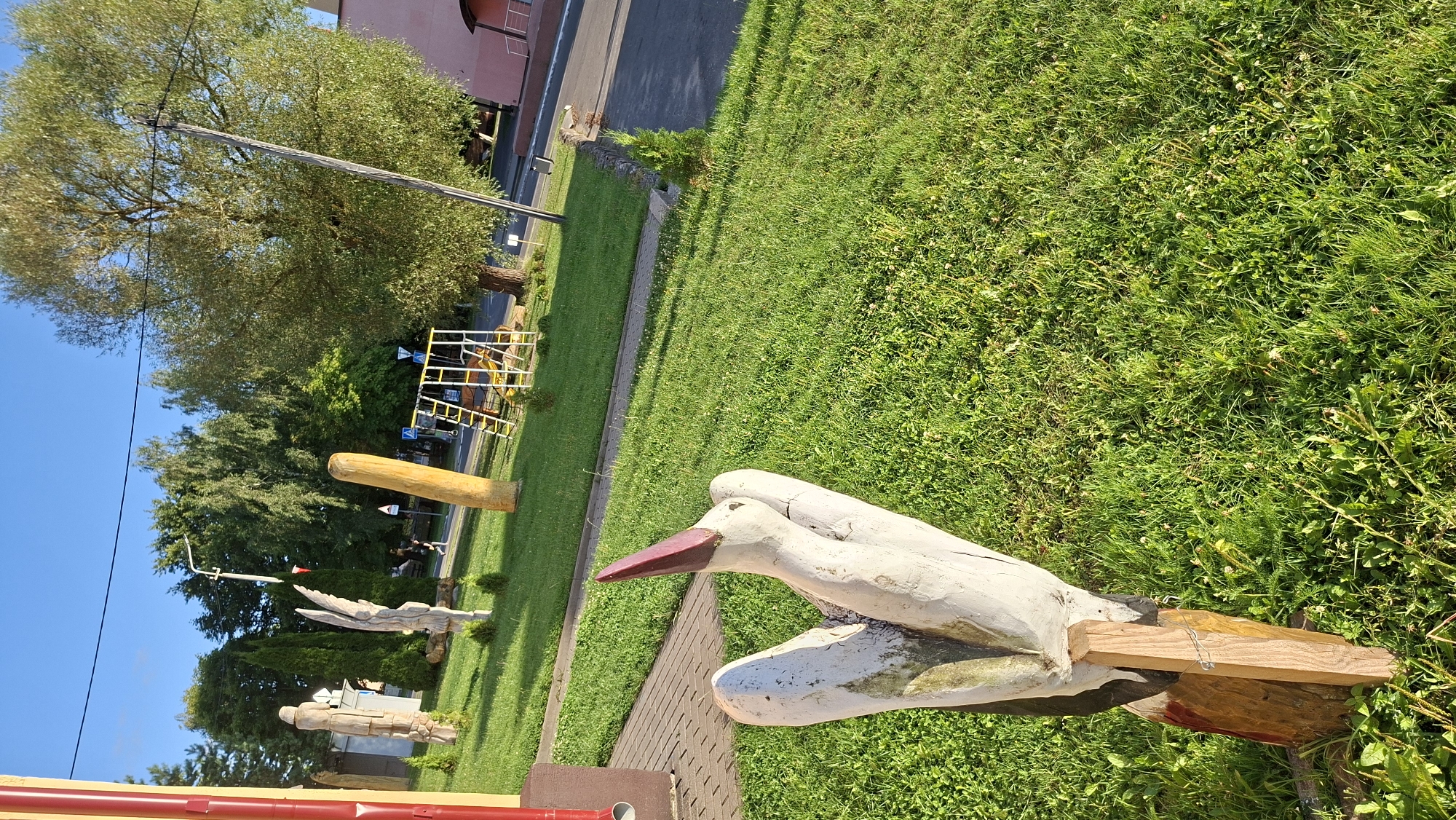 A white wooden sculpture of a stork's head and neck, with a red beak, stands in the foreground on a green lawn. The sculpture is mounted on a wooden post. In the background, there are several other wooden sculptures, including what appears to be a figure and an angel. A playground set with yellow and white bars is visible further back. Trees and a building with a pink facade are also in the background. The scene appears to be located in a public outdoor space, possibly a park or a cultural site, in Myadzyel, Belarus. The sky is blue and clear, and the sun is shining, casting shadows.