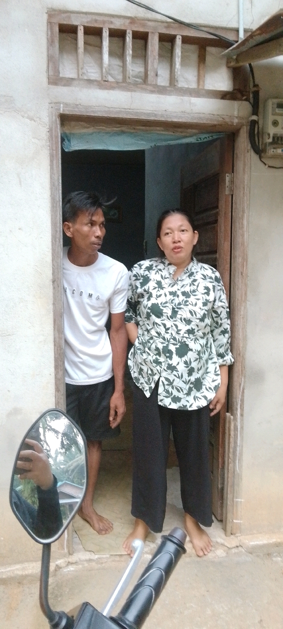 In Lenggadai Hilir, Indonesia, a man and a woman stand at the entrance of a weathered building. The man, wearing a white t-shirt and black shorts, stands to the left, looking off to the side. The woman, dressed in a patterned blouse and black pants, stands to the right, facing the camera with a neutral expression. They are positioned within a doorway that appears to be constructed of wood and has a wooden grate above it. The building's exterior wall is made of rough, light-colored concrete. In the foreground, the handlebars and side mirror of a motorcycle are visible, reflecting a person holding a phone. The ground outside the building is a concrete surface, cracked and aged. Electrical conduit and a meter box are attached to the exterior wall to the right of the doorway.