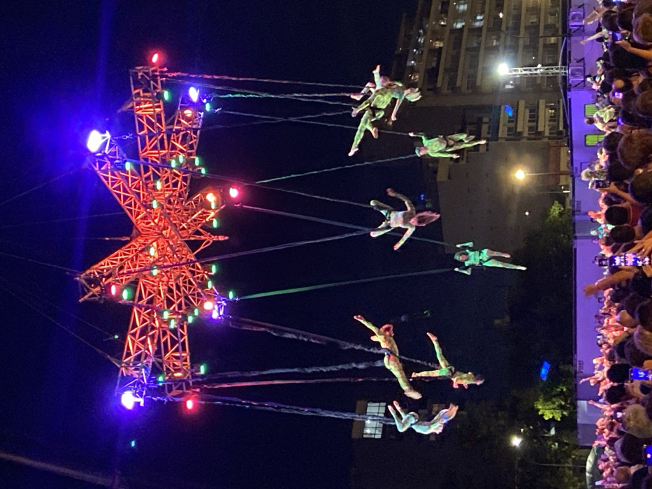At night, a group of performers suspended by wires swing from a central star-shaped structure, illuminated with red, green, and purple lights. The performers are wearing colorful outfits and appear to be floating in the air. A crowd of people is gathered to watch the performance, many holding up their phones to take pictures or videos. Tall buildings are visible in the background.