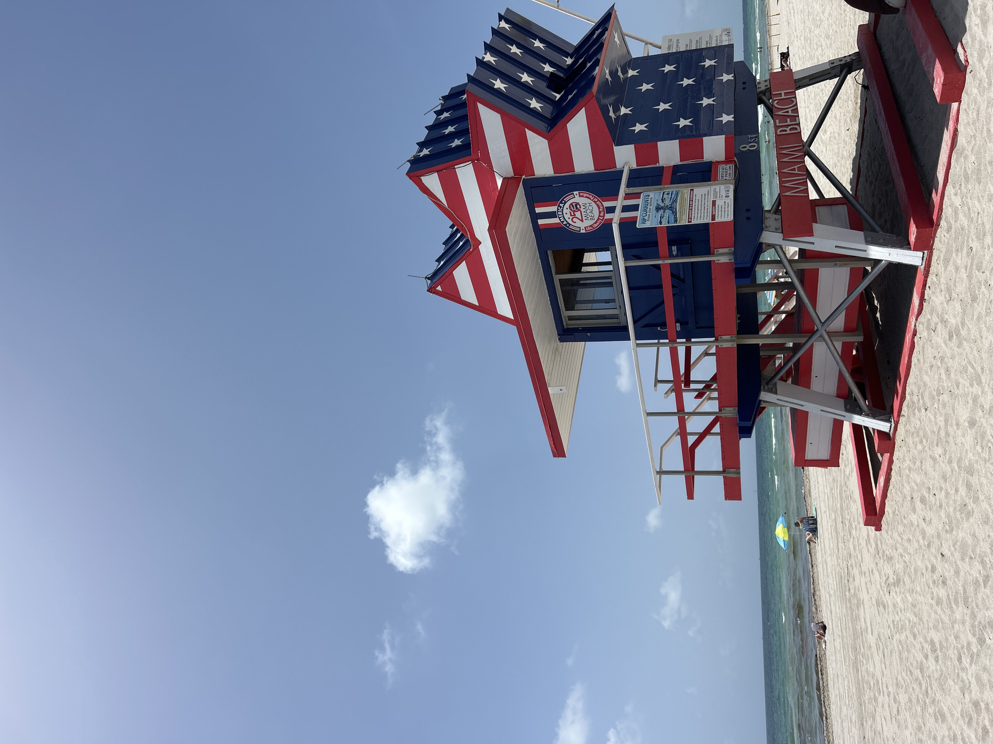 This is a vertical, eye-level, full shot of a lifeguard tower on Miami Beach. The tower is shaped like a star and is painted with red and white stripes and blue with white stars, reminiscent of the American flag. The tower has a red and white railing around a small platform. Text on the tower reads "MIAMI BEACH" and "8 ST". The background shows a calm ocean with gentle waves, a sandy beach, and a bright blue sky with scattered clouds. In the distance, two people are sitting under a yellow and blue umbrella on the beach. The overall scene conveys a patriotic and serene beach atmosphere.