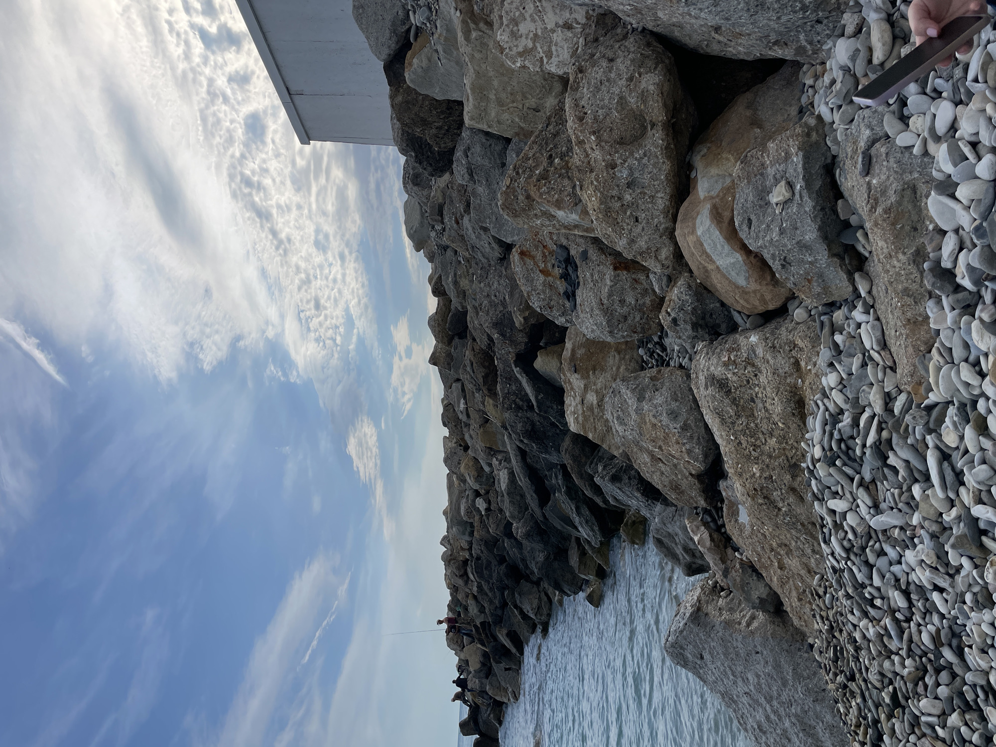 The photograph shows a view of a rocky breakwater in Olginka, Russia, on a day with a partially cloudy sky. The breakwater is constructed from large, irregularly shaped gray and brown rocks. The sea can be seen to the left of the breakwater, and a small strip of shore covered in smooth, gray pebbles is visible on the right. Several people are standing along the breakwater, some appear to be fishing with fishing rods. A white rectangular structure is visible at the top left of the breakwater. A hand is holding a black phone over the rock on the breakwater.