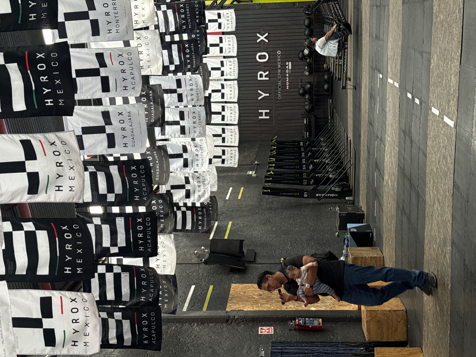 The image depicts an indoor gym or fitness facility in Mexico City, Mexico, showcasing a variety of workout equipment and branding. The scene is well-lit by artificial lights.
On the left side, numerous flags are prominently displayed, hanging vertically. These flags are predominantly black and white with red and green accents, featuring the "HYROX MEXICO" logo. Many also include the names of specific Mexican cities such as "ACAPULCO," "GUADALAJARA," and "MONTERREY." Smaller text like "FIRST PLACE," "PRO DOUBLES," and "PRO MEX 55-87" can also be seen on some flags.
In the background, a large black corrugated wall displays a prominent white "HYROX" sign, with "OFFICIAL GYM MEXICO" and "NOMADAS" written below it.
In the mid-ground, a section of the gym is equipped with several Concept 2 rowing machines, identified by their "CONCEPT 2" branding and numbers ranging from "01" to "08." Racks of medicine balls are also visible. One man, wearing a white t-shirt and dark shorts, is actively engaged in exercising on a rowing machine. Further to the right, another man, dressed in a black t-shirt and blue jeans, stands holding a baby. He is looking down at a smartphone in his hand. The baby is wearing a striped outfit.
The floor is covered with dark rubber gym mats featuring contrasting stripes and white and yellow marking lines. Wooden boxes are also placed on the floor. The overall branding suggests an official Hyrox competition or training facility.