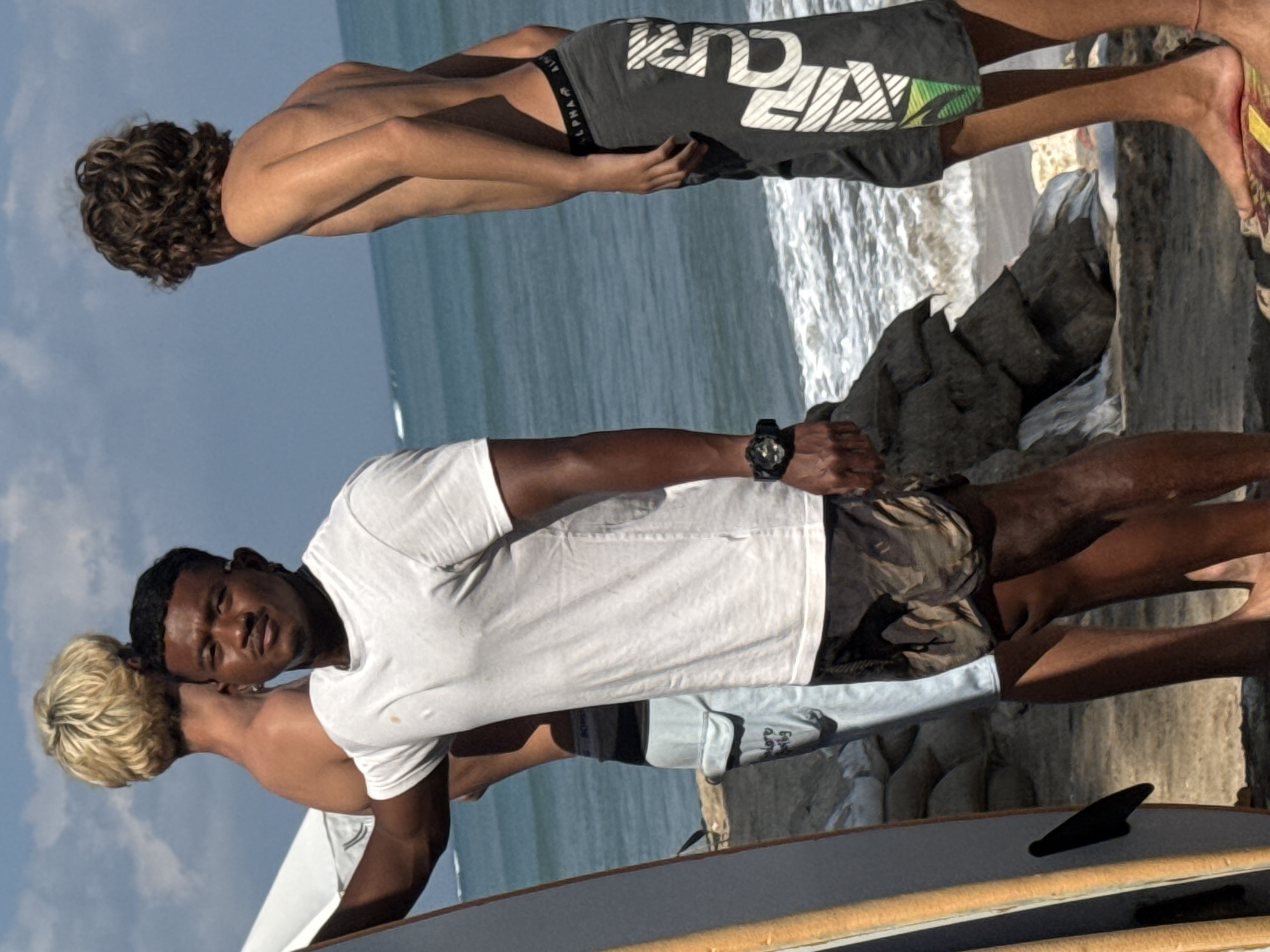 In Kuta, Indonesia, on a sandy beach with a backdrop of the ocean, three young men stand near a surfboard. One man, of darker complexion, in a white t-shirt and camouflage shorts, faces the camera, a serious expression on his face. He wears a black digital watch on his left wrist. To his left, another man with blonde hair, back to the camera, wears light blue shorts. To the right, a third man with curly brown hair, also with his back to the camera, wears dark grey shorts with a white graphic design and the word "ALPHA" visible on the waistband. The ocean, with gentle waves, stretches to the horizon under a clear sky. Sandbags are visible in the background, suggesting the area might be prone to erosion or flooding. A surfboard, leaning against the man in the white shirt, is partially visible on the left side of the frame.