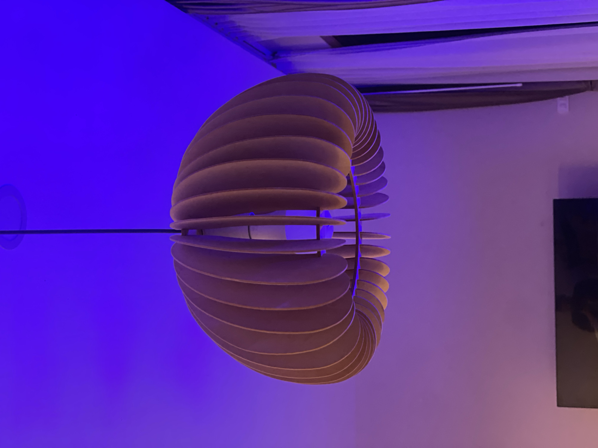 A wooden pendant light fixture hangs from the ceiling in a room with purple ambient lighting. The fixture is composed of numerous curved wooden slats arranged in a spherical shape. To the right of the fixture are light-colored curtains. The bottom of the image shows the top of a black television screen. The scene is indoors, and it is not possible to determine the specific location within Baan Plai Laem, Thailand.