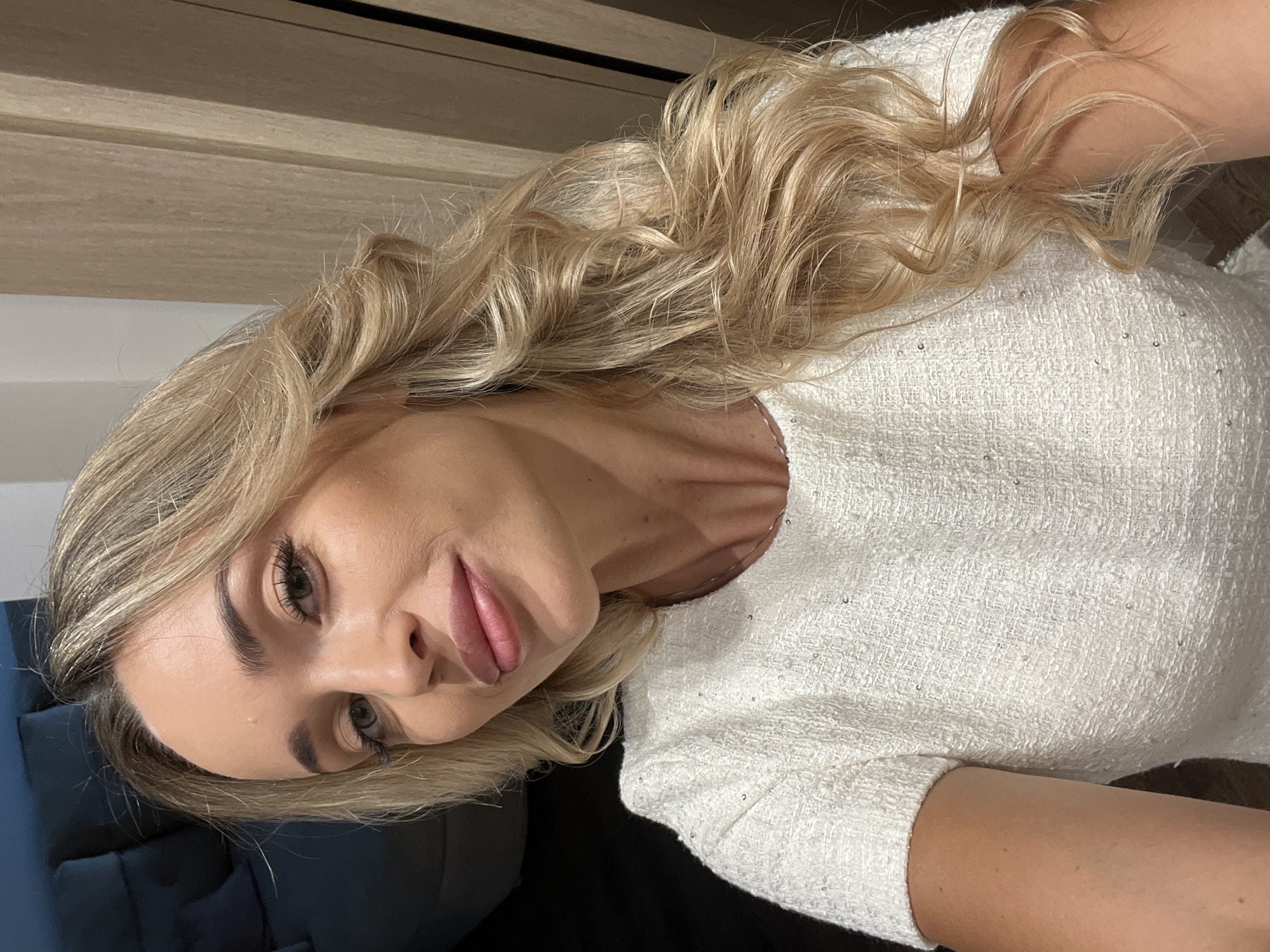 A blonde woman with wavy hair is taking a selfie. She is wearing a white textured dress with short sleeves. Her hair is parted to the side and falls over her right shoulder. She is looking towards the camera with a slight smile. Her makeup is natural, with emphasis on her eyes and lips. The background appears to be indoors, with a wooden closet or door visible to the right and a dark sofa to the left. There is no indication of the location being Sharm El Sheikh, Egypt.