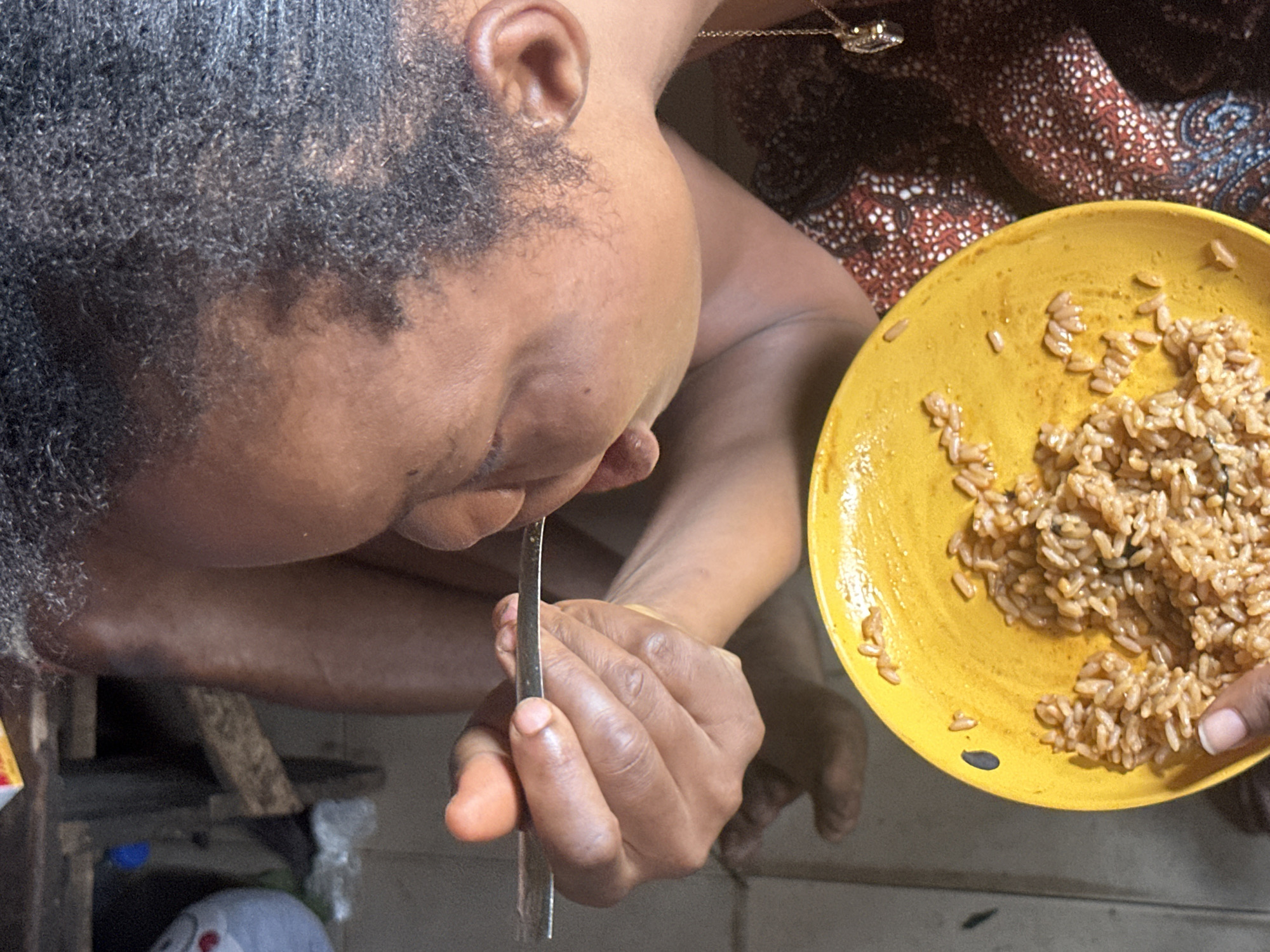 A person with dark skin, curly hair, and a gold necklace is shown eating rice from a yellow plate with a spoon. The individual is seated indoors, possibly in a home in Aba, Nigeria. The environment appears to be dimly lit with basic wooden furniture visible in the background. The plate contains a significant portion of cooked rice, and the individual is in the process of bringing a spoonful to their mouth. Their focus is on the act of eating. A patterned brown, white, and blue cloth is draped over their shoulder.