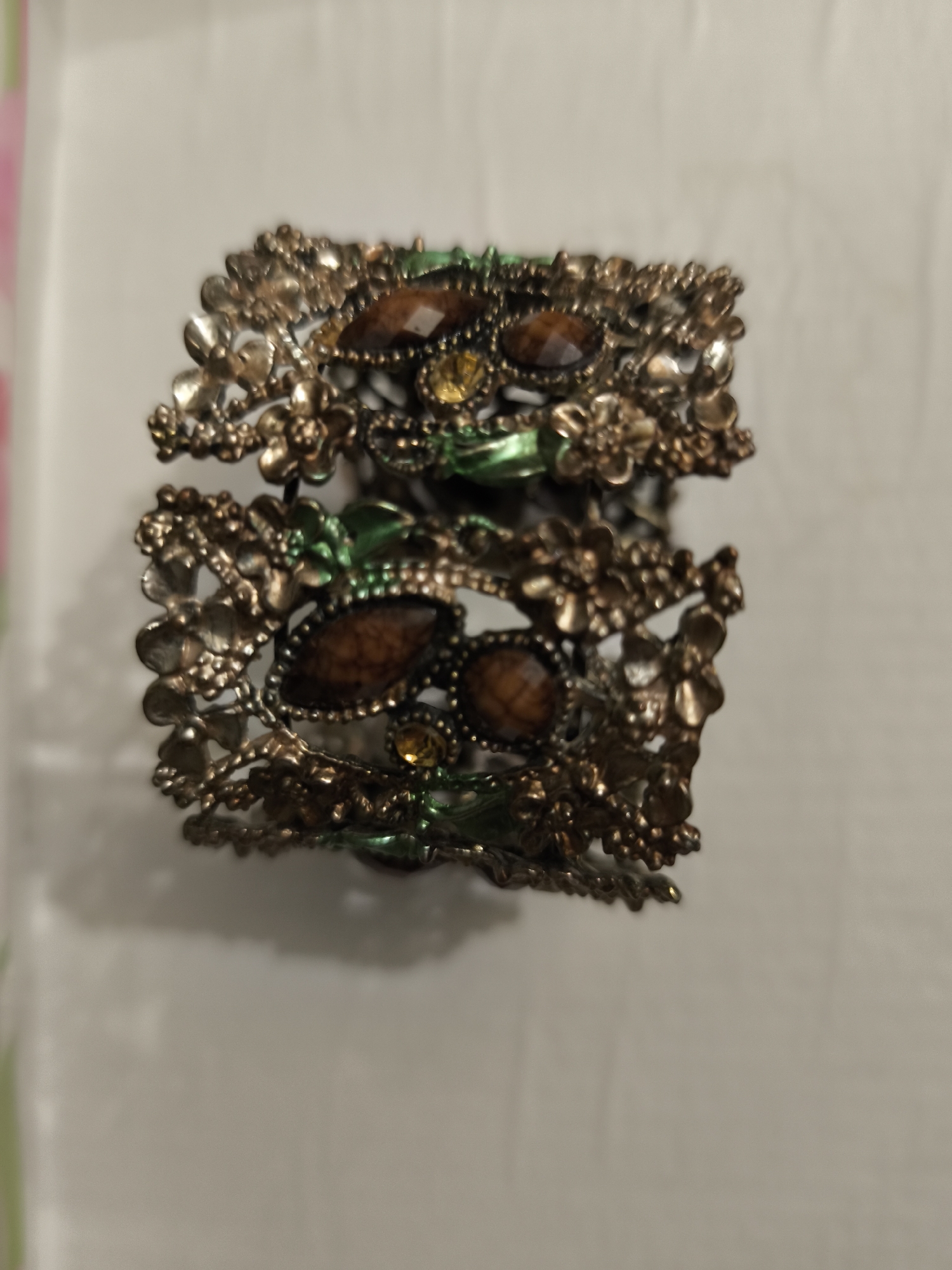 A close-up, slightly blurry shot shows an ornate, bronze-colored metal cuff bracelet with floral motifs and inset brown and yellow gemstones. The bracelet is designed in two tiers, with the upper tier featuring a more prominent arrangement of flowers and gemstones, while the lower tier is more delicate. The background is a plain, light-colored surface, possibly a table or wall, with a soft shadow cast by the bracelet. There is no indication of the bracelet's location in the city of Bolshaya Martynovka, Russia, as the image is an isolated product shot.