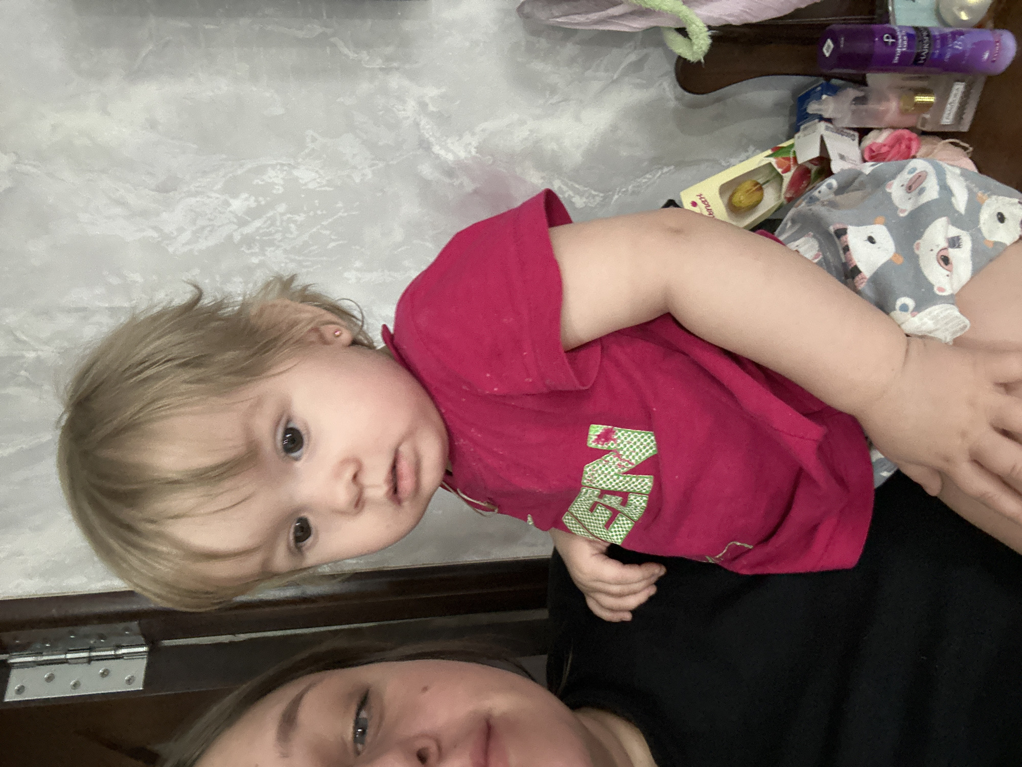 The image shows a child wearing a red shirt with green lettering lying on top of a person wearing a black shirt. The background includes a textured wall and an assortment of items on a surface, including toiletries and boxes. The location is Ольховка, Russia.