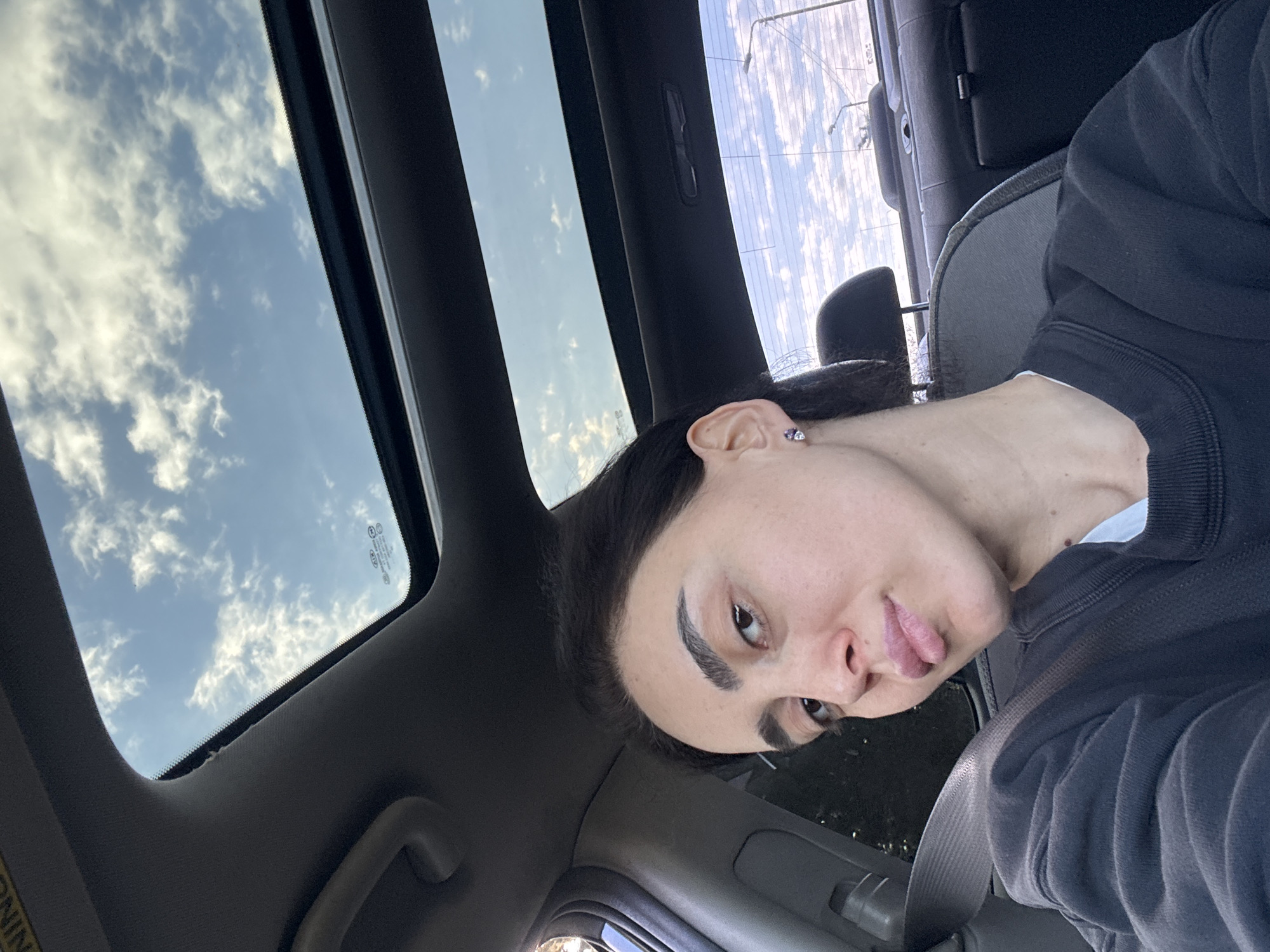 A person is sitting in a car with an open sunroof. Outside the sunroof, the sky is visible with scattered clouds. The person has dark hair, and the interior of the car is dark gray or black. The person is wearing a dark-colored sweater. The seatbelt is visible. The location is not evident in the image.