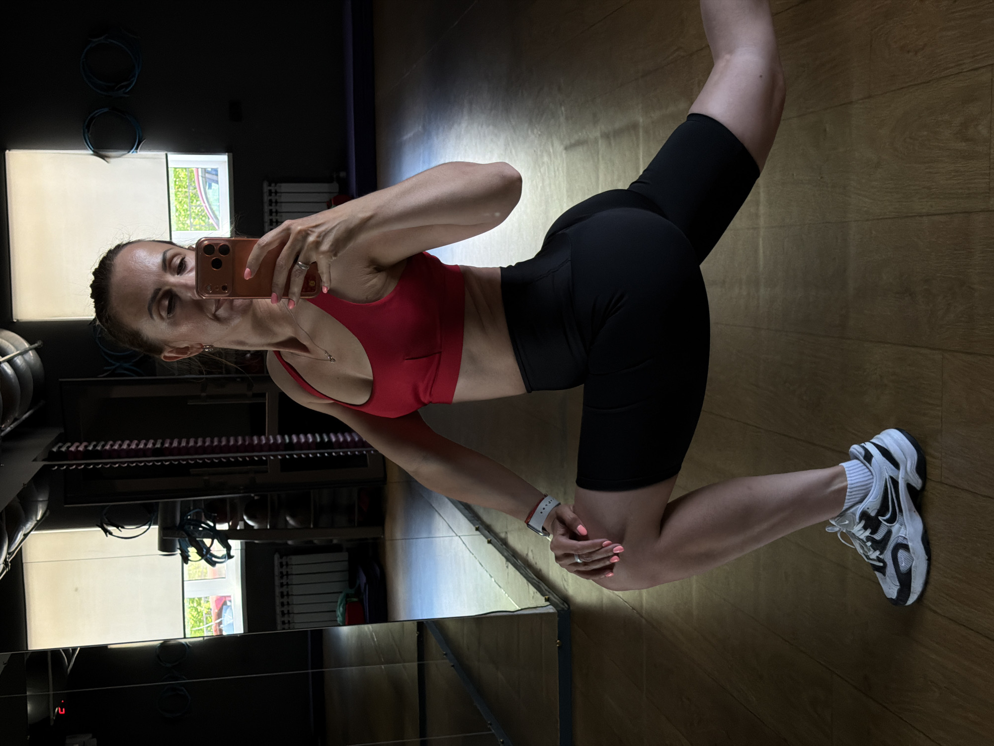 The image features a woman taking a mirror selfie in what appears to be a gym. She is wearing a red sports bra and black biker shorts. Her hair is pulled back in a ponytail, and she is wearing a watch on her left wrist and a ring on her right ring finger. Her fingernails are painted pink. She is holding a rose gold smartphone in her right hand, which she is using to take the picture.
The setting is a gym with wooden flooring. In the background, there are gym equipment, including what looks like weight racks and resistance bands. There is also a large mirror reflecting the scene. Through a window in the background, greenery can be seen, suggesting it is daytime. The lighting in the gym is somewhat dim, with some areas brightly lit and others in shadow. The overall mood is casual and athletic. There is no visible text in the image. The location is Birobidzhan, Russia.