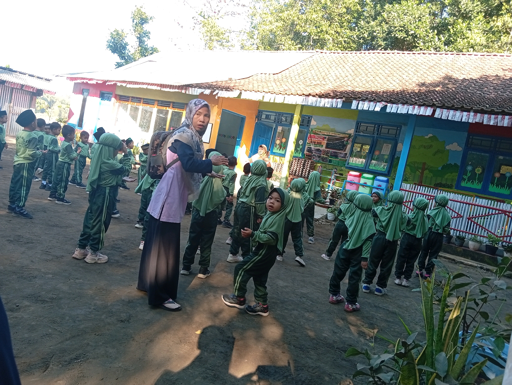 In Miritpetikusan, Indonesia, on a sunny day, a group of young children, all wearing green uniforms with headscarves, are gathered outdoors in front of a brightly painted school building. The building features colorful murals depicting trees and flowers, with blue-framed windows. A woman, also wearing a headscarf and a light-colored tunic over dark pants, stands among the children, seemingly instructing or supervising them. Some children are looking at the camera, while others are engaged in activities, with one child in the foreground performing a leg-raising exercise. The ground is dusty and uneven. The scene appears to be a schoolyard during an outdoor activity or lesson.