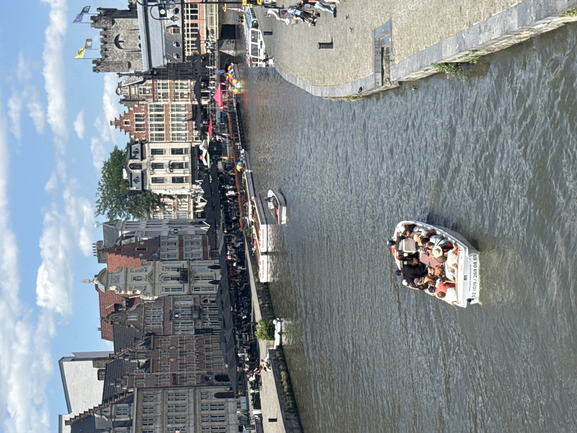This image depicts a vibrant scene in Ghent, Belgium, with a canal running through the city. The time of day appears to be daytime, with clear blue skies and scattered clouds, suggesting pleasant weather. The architecture along the canal is characteristic of Ghent, featuring historic brick buildings with distinct gabled roofs and large windows.
Several boats are visible on the canal. The most prominent is a white tour boat filled with people enjoying a scenic ride. Passengers are seated, some wearing hats, and appear relaxed and engaged with their surroundings. The boat has visible text on its side: "32 (0)9 / 269 08 69", likely a phone number for boat tours. Another smaller white boat is also present.
Along the canal's edge, there's a cobblestone walkway lined with outdoor seating areas, indicating a popular spot for cafes or restaurants. People are gathered at these tables, some under black umbrellas, suggesting a lively atmosphere. Further along the canal, a procession of colorful flags, including rainbow-colored ones, adds a festive touch to the scene. The overall mood is one of a bustling, enjoyable day in a historic European city.