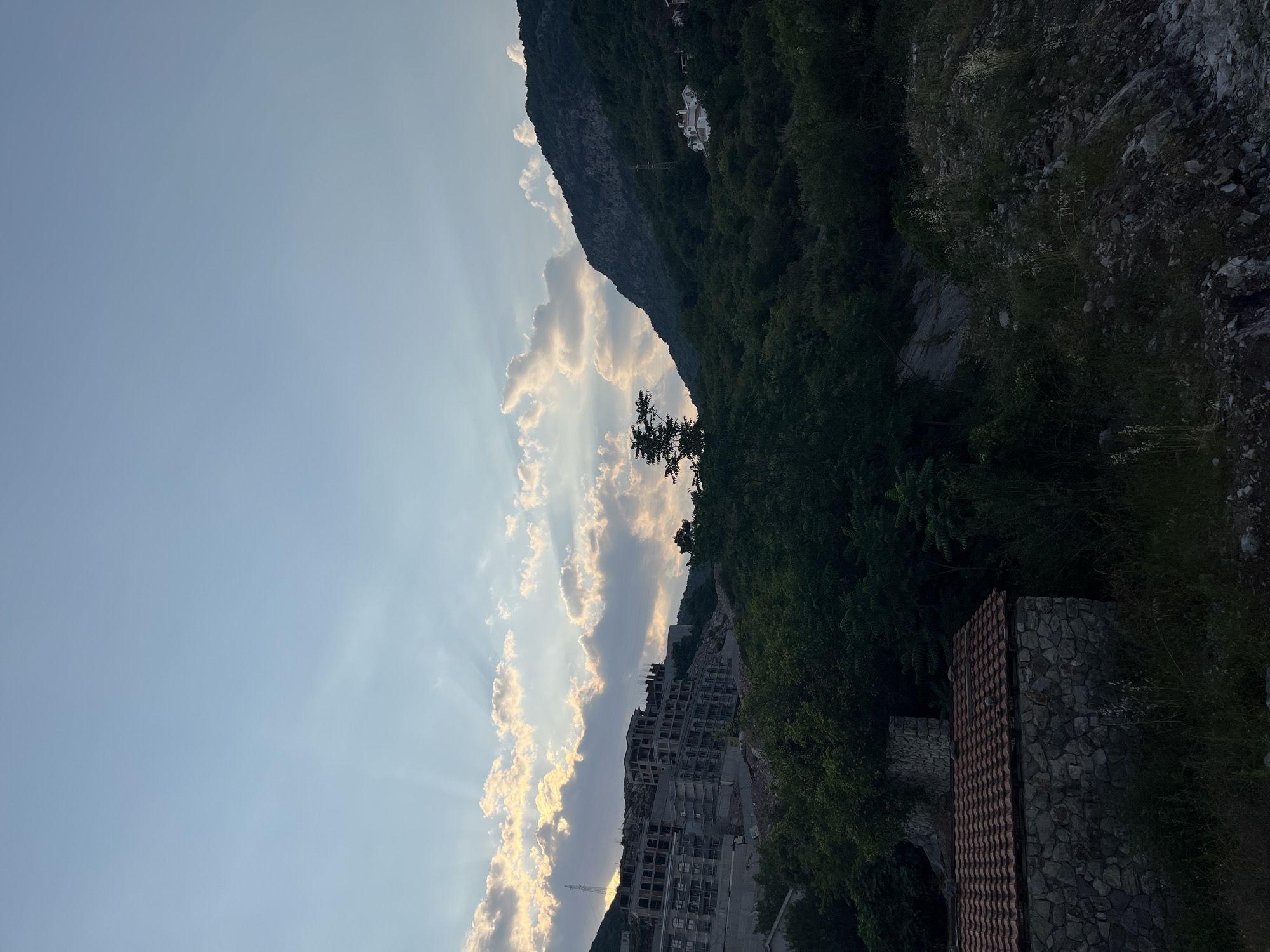 The image captures a dramatic landscape scene in Budva, Montenegro. The primary subjects are a steep, verdant mountainside and a vast sky filled with dramatic clouds. Below the mountain, a partially constructed, multi-story building is visible, its concrete structure hinting at a stalled development. The foreground features a weathered stone structure with a tiled roof, adding a rustic element to the scene.
The time of day appears to be late afternoon or early evening, indicated by the soft, angled sunlight filtering through the clouds and casting long rays. The weather seems clear, with the sun breaking through scattered clouds, creating a beautiful, ethereal light.
Notable details include the dense green foliage covering the mountainside, contrasting with the rocky texture of the upper mountain slopes. The unfinished building suggests a complex or perhaps abandoned project. The stone building in the foreground, with its traditional tiled roof, provides a sense of history or local architecture. The overall mood is serene and awe-inspiring, with the natural beauty of the mountains and sky dominating the composition.
No visible text is present in the image. The image is oriented vertically.