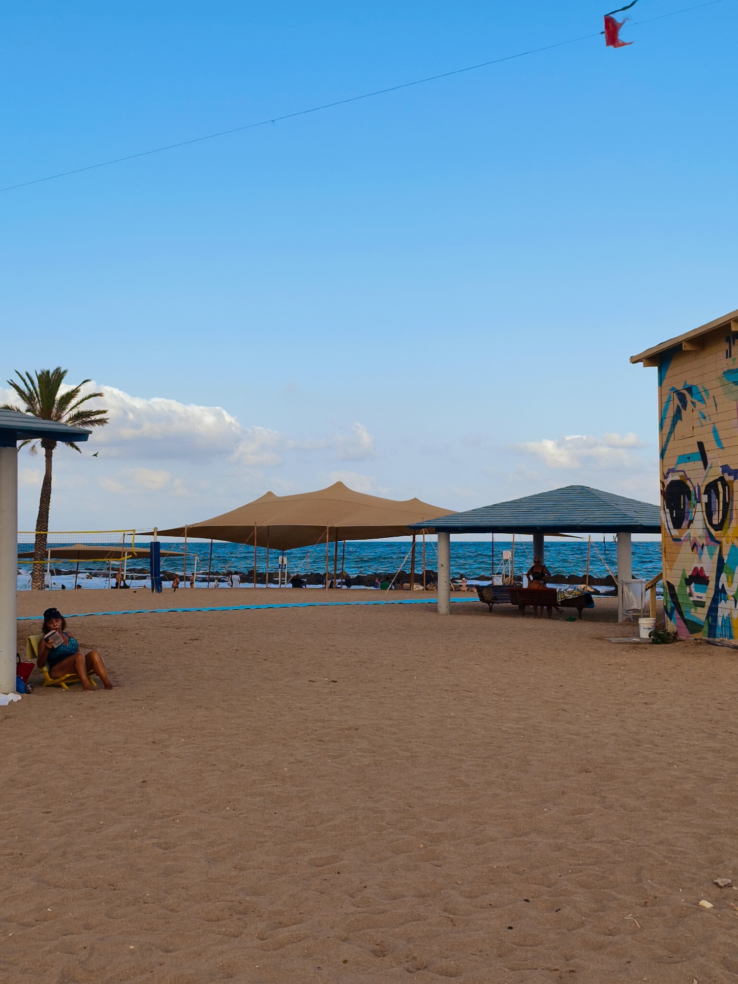 This image depicts a beach scene in Nahariya, Israel, on a clear day with a few clouds. The foreground is dominated by a sandy beach. In the midground, the sea is visible, with waves breaking against a rocky shoreline. Several beach amenities are present, including large tan and blue canopies providing shade, volleyball nets, and a palm tree.
On the left side of the image, a woman is seated on a yellow lounge chair, reading a book. She is wearing a blue bikini and a dark cap. Further back, under the blue canopy, a person is seated on a bench.
To the right, a wooden building is adorned with colorful graffiti, featuring a large stylized face with prominent glasses. A white bucket is placed near the building.
In the sky, a thin wire stretches across, and a small red object, possibly a kite or flag, is attached to it. A bird is also visible in flight. The overall impression is of a relaxed and vibrant beach atmosphere.