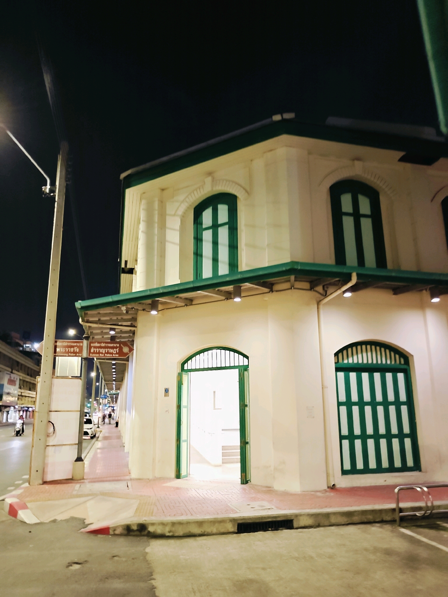 This is a nighttime, medium-distance, eye-level shot of a building with a cream-colored exterior and green trim, located in Bangkok, Thailand. The building has a colonial architectural style and features arched windows and doors with green shutters. The entrance to the building is a large, arched doorway with an open green gate, revealing a bright interior. To the left of the building, a sidewalk runs along a street with traffic and streetlights. A sign on a pole indicates a police area. The sky is dark, and the scene is illuminated by streetlights and lights from the building and the street. There are no people visible in the immediate foreground, but several figures can be seen walking on the sidewalk in the distance. The overall impression is of a quiet street scene at night in a historical part of the city.
