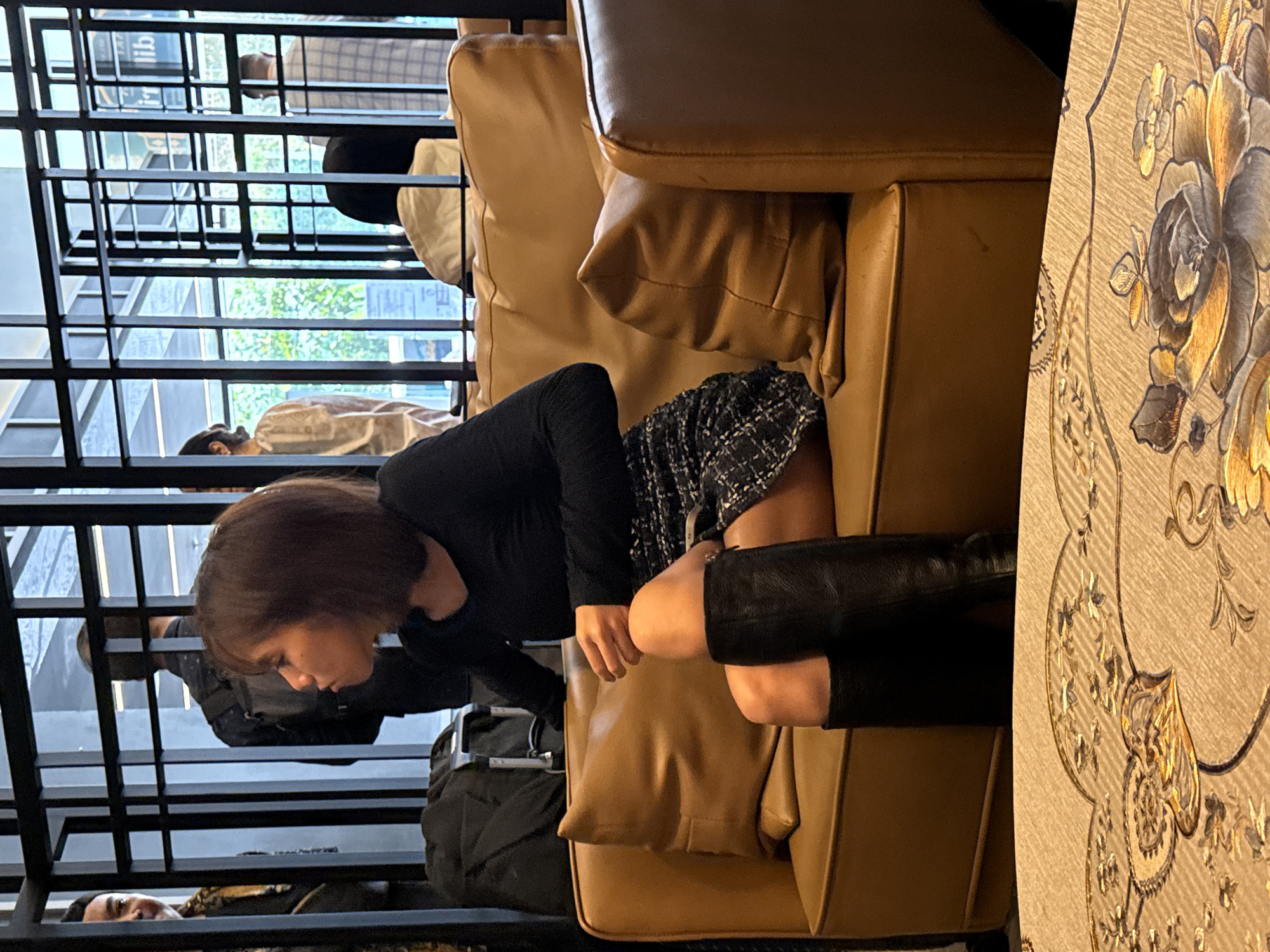 The image captures a woman seated on a tan leather chair or sofa. She is wearing a black long-sleeved top, a tweed skirt, and black leather boots. She is in what appears to be a waiting area or lounge, as there are black metal bars visible in the background, possibly from a structure or enclosure, and other people are also present in the distance. Based on the visual information, it is not possible to confirm if the location is in Kuala Lumpur, Malaysia.