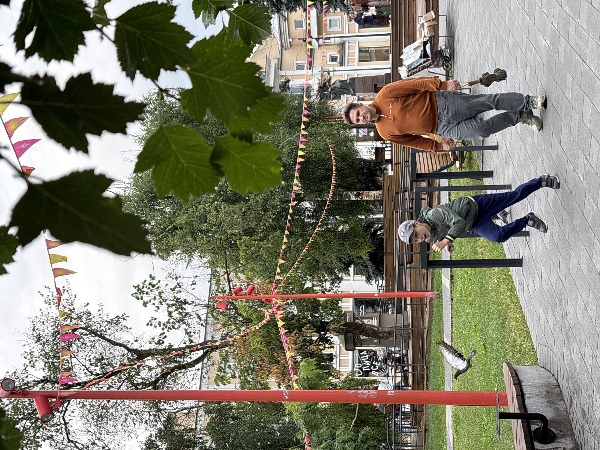 In Nizhny Novgorod, Russia, on an overcast day, a boy in a grey cap and green jacket runs towards a pigeon taking flight. Behind him, a man in an orange sweater and grey pants walks with an umbrella. Banners hang from red poles, decorating the outdoor area. The background reveals buildings, trees, and people observing the scene.