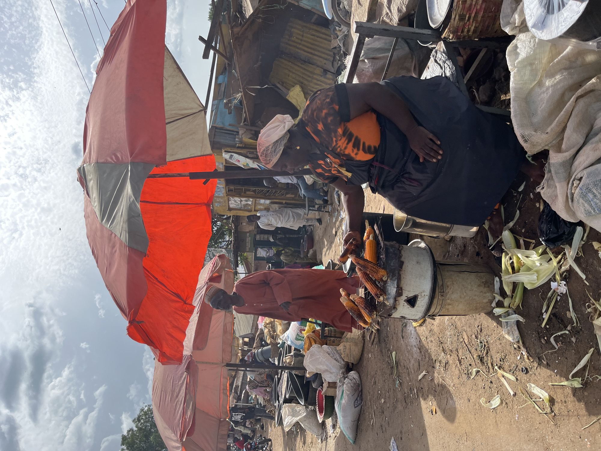 A bustling outdoor market scene in Jos, Nigeria, is depicted under a cloudy sky. The foreground features a woman in a black and orange tie-dye shirt and a dark blue apron, attending to a roasting apparatus that holds several ears of corn. She wears a light-colored head covering. Beside her, a man in a rust-colored robe and a dark cap stands, appearing to interact with the corn roaster.
The market is filled with various stalls and goods. Large, colorful umbrellas, predominantly orange and red, provide shade. Numerous sacks, some appearing to contain produce or grains, are piled around the area. Behind the main subjects, the market continues with more stalls, structures made of corrugated metal, and other people moving about. In the background, a few motorcycles are visible, suggesting transportation options. The ground is dirt, with scattered corn husks and debris, indicating a busy, working market environment. The lighting suggests it is daytime, possibly late morning or early afternoon, with diffused sunlight due to the clouds. The overall atmosphere appears to be one of active commerce and daily life in a Nigerian marketplace. No visible text is discernible.