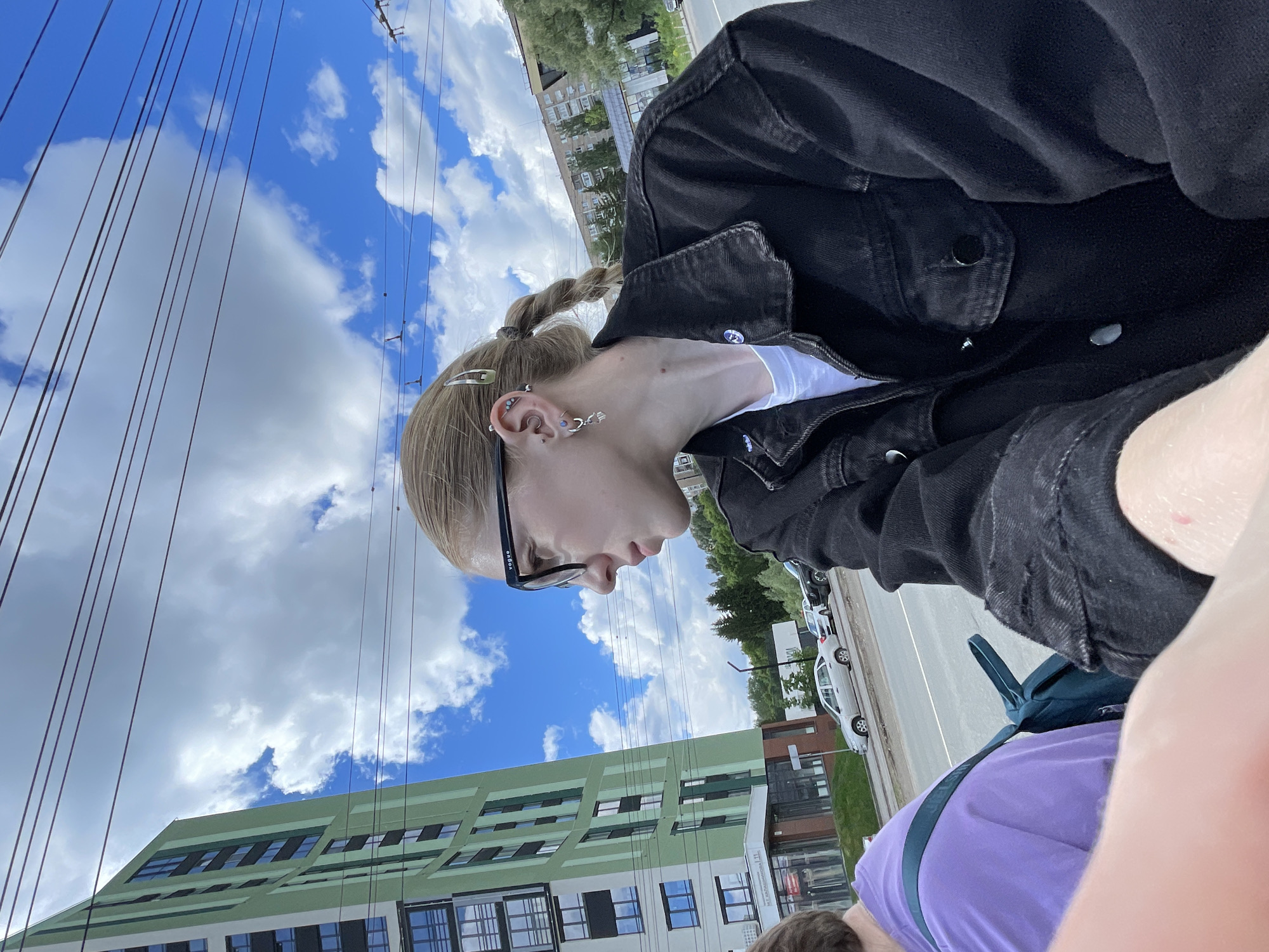 A young woman in a black denim jacket and glasses is shown in a medium shot, presumably a selfie. She has fair skin and blonde hair styled in a short braid. Several piercings adorn her ear. The background reveals an urban environment. A building with green and white panels and numerous windows is visible. Overhead electrical wires crisscross the sky, which is filled with scattered white clouds. A road with parked cars lines the background. The location is identified as Izhevsk, Russia.