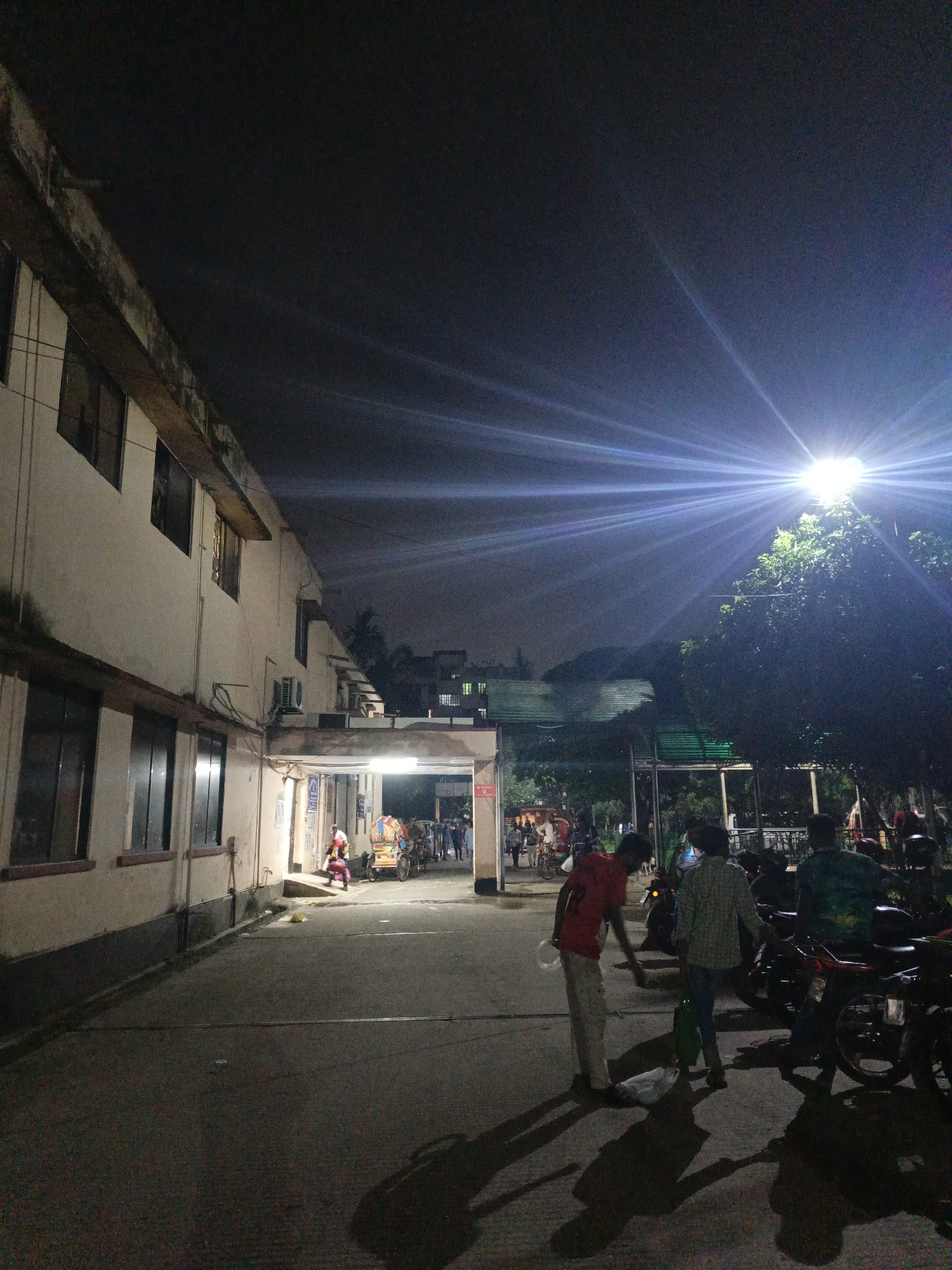 At night in an urban environment of Unknown, Unknown, a two-story building stands to the left of the frame. A single person walks under a covered walkway. In the middle of the frame, two men walk down a street, and a group of people congregate further down the street. To the right, a row of motorcycles sit parked. An overhead light shines down on the scene.