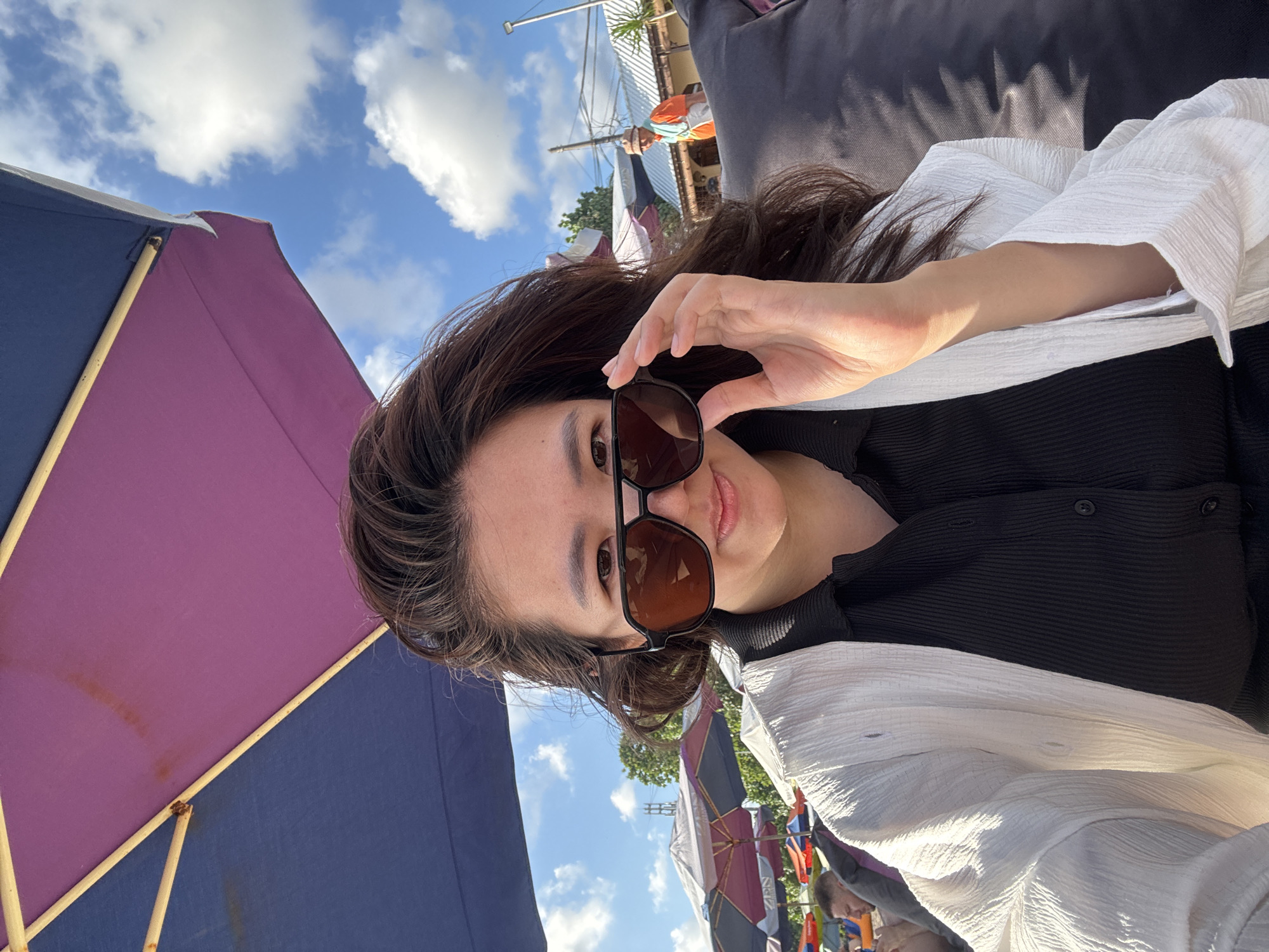 A woman with dark hair is taking a selfie at a beach in Kuta, Indonesia. She is wearing sunglasses and a white cover-up over a black shirt. In the background, there are beach umbrellas, other people, and buildings. The sky is blue with white clouds.