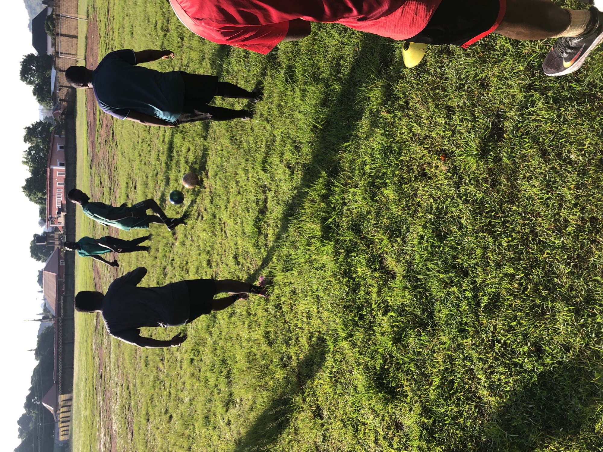 The image depicts a group of young men practicing soccer on a grassy field in Jos, Nigeria. The scene is set in an open area with a soccer field, characterized by green grass and dirt patches. In the background, there are buildings with red roofs, a fence, and trees, suggesting an urban or suburban environment. The lighting indicates it is either early morning or late afternoon, with long shadows cast across the field.
There are at least five individuals visible. One person in the foreground is wearing a red t-shirt and black shorts, with a yellow soccer cone near their feet. Their back is to the camera. Another person, in the center-left, is wearing a dark grey t-shirt and black shorts and is captured from behind, appearing to be running. To the left of this individual, another person in a green shirt and black shorts is also facing away from the camera, with one foot on a soccer ball. Further back and to the left, a person in a green shirt is visible from the side, appearing to be in motion. On the right side of the image, a person in a teal or dark blue shirt and black shorts is also visible from behind.
The men are engaged in a soccer training session, with two soccer balls clearly visible on the ground. The overall atmosphere suggests an active and athletic setting. The presence of soccer balls, cones, and the grassy field clearly identifies the activity. The backdrop with buildings and trees places the scene within a developed area in Jos, Nigeria, likely a sports field or training ground.