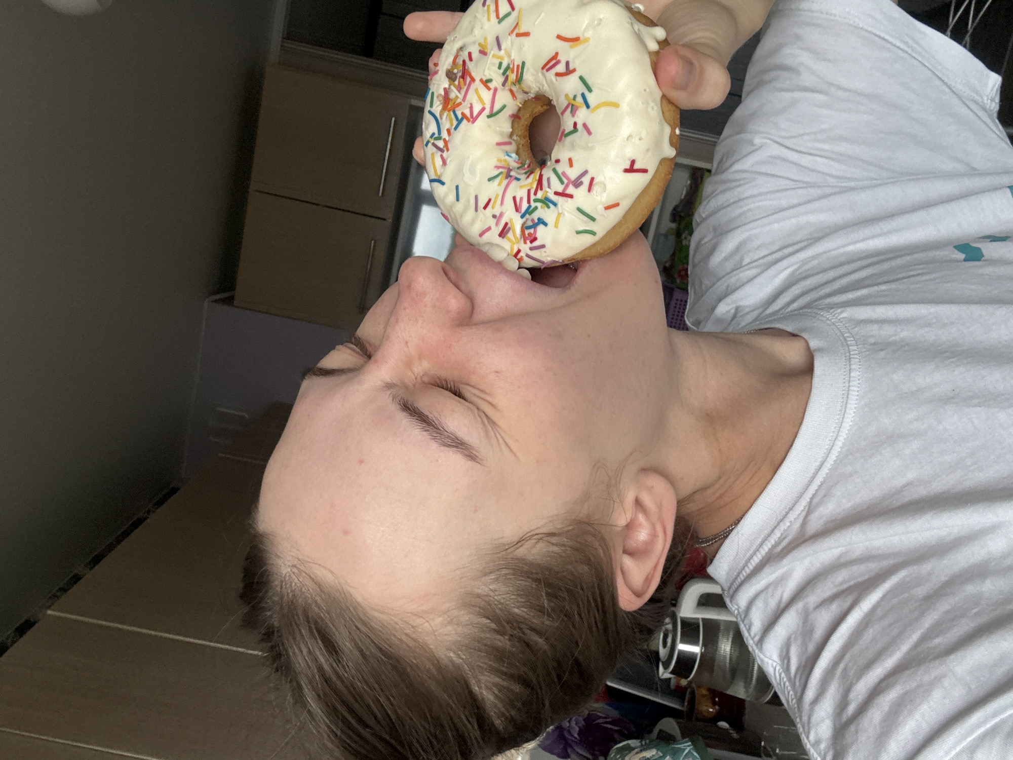 A young woman is biting into a white frosted donut with rainbow sprinkles. She is wearing a white t-shirt. The background appears to be a kitchen with cabinets and a kettle visible. There is no information available to determine the location in the city of Perm, Russia.