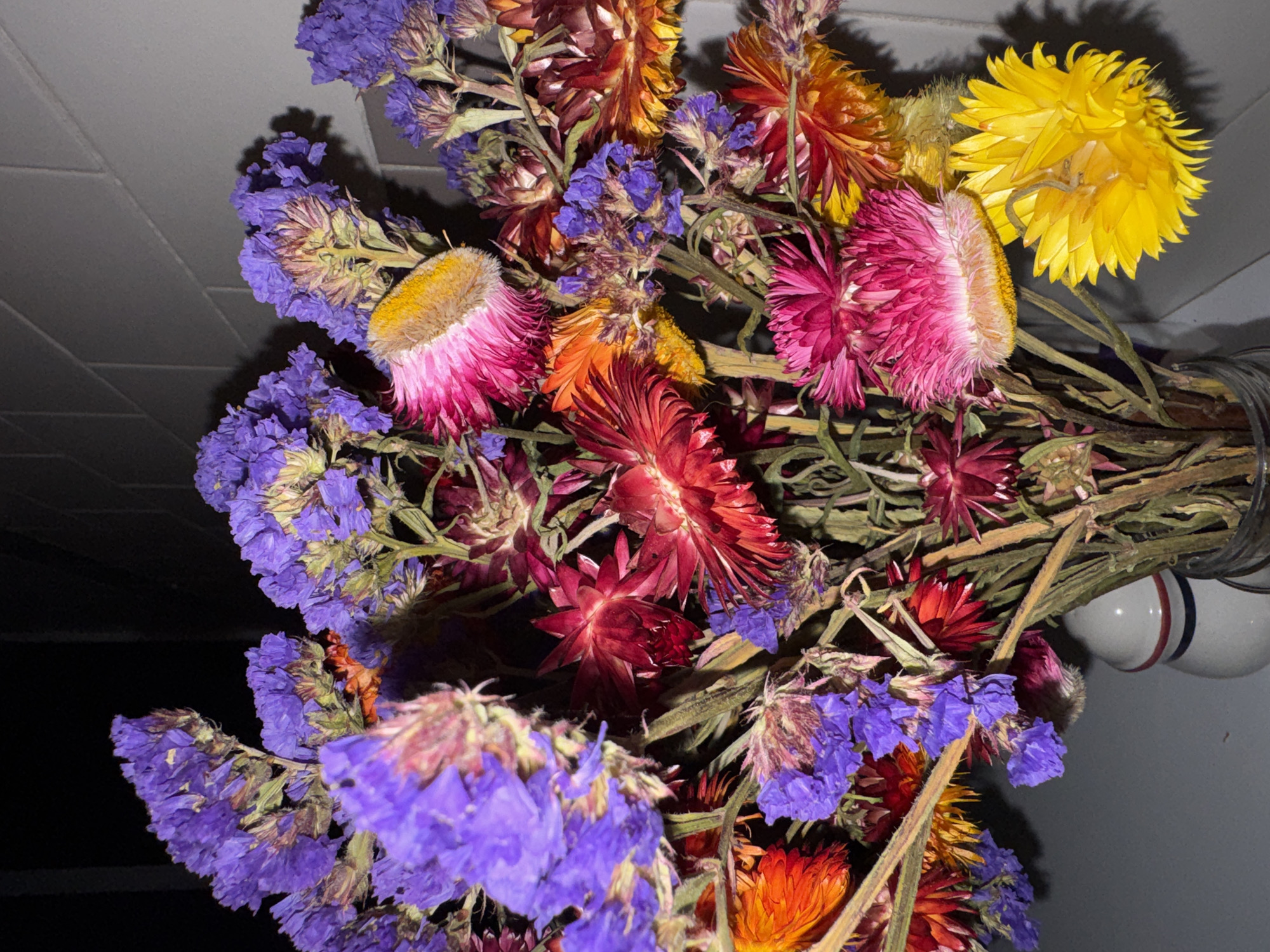 A close-up shot of a bouquet of dried flowers. The flowers are a mix of purple, pink, yellow, orange, and red. The stems are brown and dry. The bouquet is in a vase. The background is a dark gray wall. The location is not specified.