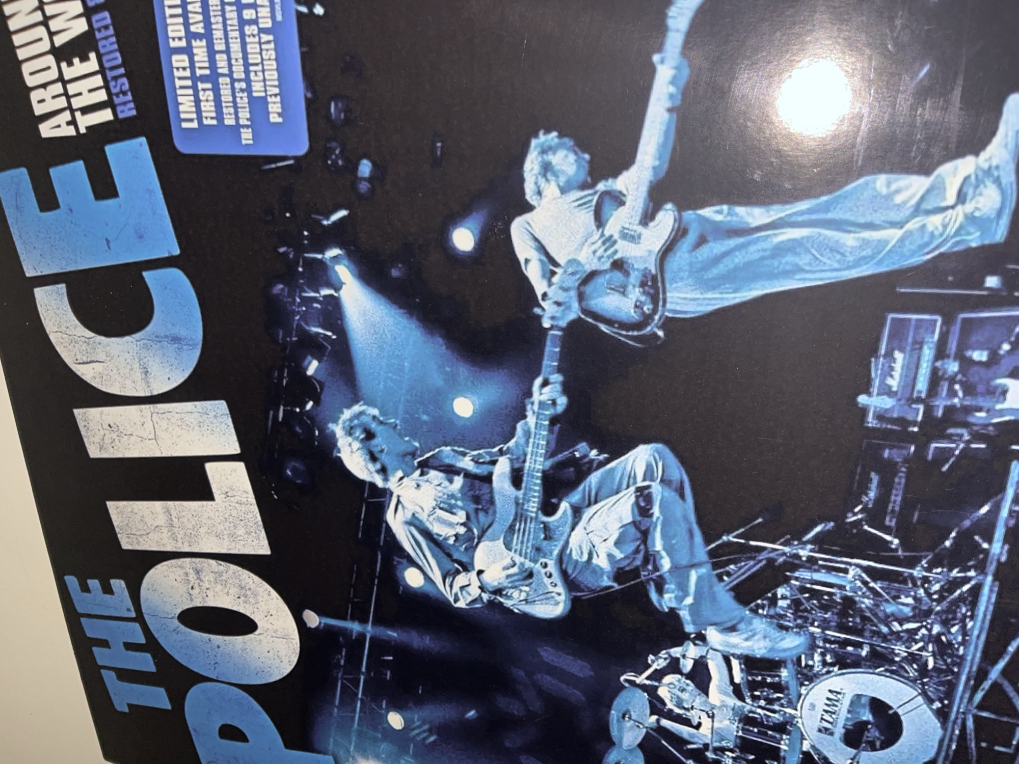 An image, likely a album or DVD cover, displays a dynamic, dark-toned scene of a live concert performance, predominantly in shades of blue and black. Two musicians are the central figures. One musician, positioned higher and appearing to jump or float mid-air, plays an electric guitar with his legs extended. Below him, another musician with spiky hair is crouched or seated on equipment, actively playing a bass guitar.
The setting is a dimly lit stage, illuminated by bright blue stage lights creating stark contrasts and shadows. Visible stage equipment includes parts of a drum kit, specifically a bass drum with the "TAMA" logo, and what appears to be a "Marshall" amplifier stack on the right side. The overall mood is one of high energy and intense performance.
The image is cropped and rotated, with text overlaid on the left side. Large, stylized white and blue text reads "THE POLICE". Above this, partially visible text includes "AROUND THE W" and "RESTORED". A smaller blue rectangular box contains further details: "LIMITED EDITI", "FIRST TIME AVAL", "RESTORED AND REMASTER", "THE POLICE'S DOCUMENTARY", "INCLUDES 9", and "PREVIOUSLY UNA". This text suggests the media file is related to a re-released or special edition concert film or documentary by the band The Police. No specific location cues for Torrent, Spain are visible in the image.