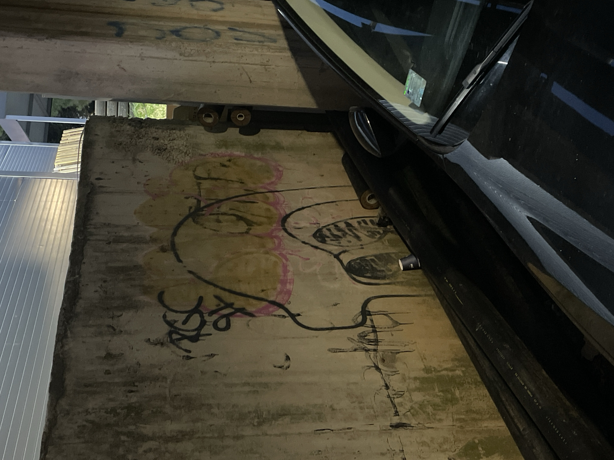 The image depicts a concrete wall with graffiti and a portion of a black car. The graffiti on the wall includes a cartoonish character with a large head and pink and yellow coloring, as well as black spray-painted tags. The wall appears weathered and stained in places. To the right of the wall, a black car is visible, with its hood and windshield in the foreground. The car has a windshield wiper and what appears to be a sticker on the windshield. A stack of dark, cylindrical objects is positioned against the wall. The scene is dimly lit, suggesting it may be nighttime or an indoor area with limited light. The overall impression is an urban or industrial setting.