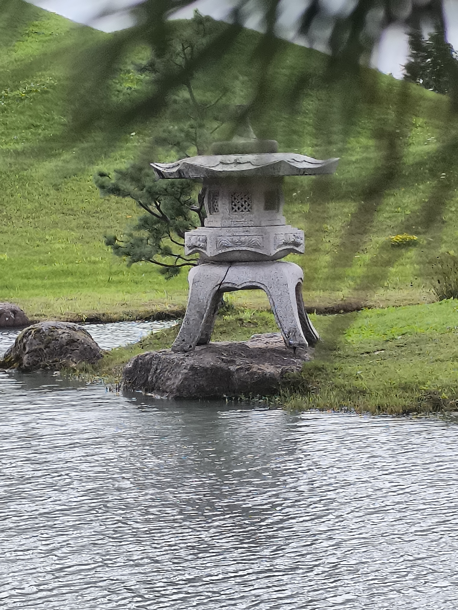 A stone lantern sits on a rock formation beside a body of water. The lantern is traditional in design with intricate carvings. The water is rippling, suggesting a light breeze. The background consists of lush green grass and trees. The overall scene appears to be a serene outdoor setting, possibly a park or garden in Mažučiai, Lithuania.