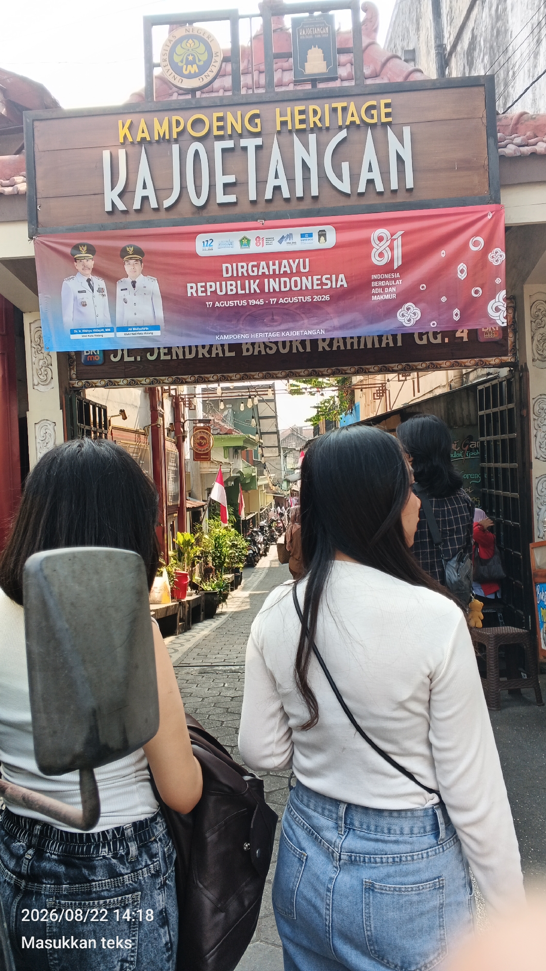 The image captures an outdoor scene at Kampoeng Heritage Kajotangan in Malang, Indonesia. A large wooden sign with the text "KAPOENG HERITAGE KAJOETANGAN" is prominently displayed above an entrance. Below this, a red banner features images of two men in official attire, likely government officials, and text that reads "DIRGAHAYU REPUBLIK INDONESIA 17 AGUSTUS 1945 - 17 AGUSTUS 2026." Further down, another sign reads "JL. JENDRAL BASUKI RAHMAT GG. 4".
In the foreground, two individuals are seen from behind, walking into the heritage area. The person on the left wears a white tank top and distressed denim jeans, carrying a dark brown jacket over their shoulder. The person on the right wears a long-sleeved white top and blue jeans. The ground is paved with cobblestones, and the street is lined with buildings and potted plants. Several motorcycles are parked along the street, suggesting it is a public area accessible by vehicle. The overall impression is of a bustling, culturally significant location within the city of Malang.