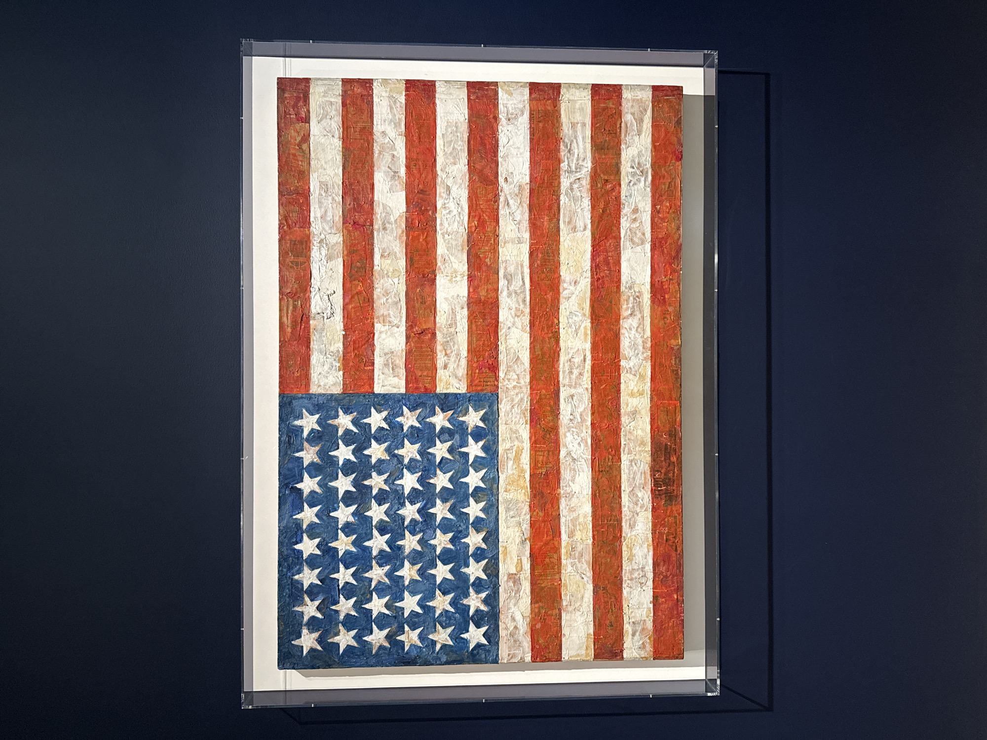 An oil painting of the United States of America flag in a clear, acrylic frame hangs on a dark blue wall. The painting, in the style of Jasper Johns, features red and white stripes and a blue field with white stars. The texture of the paint is thick and heavily applied. The location is likely a museum or gallery in New York City, USA.