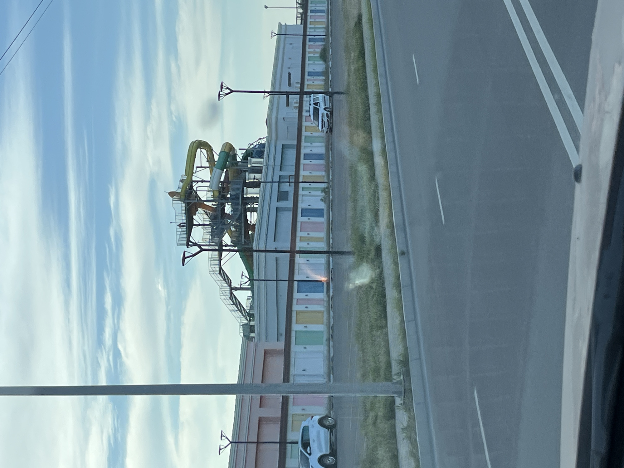 The image captures a scene from Aktau, Kazakhstan, featuring a water park in the background. The foreground shows a paved road with white lane markings. To the left of the road, a grassy area is visible, bordered by a curb. A tall, grey pole stands prominently in the left half of the image, running vertically. Behind the grassy area, a long, low building with a pink facade and a row of uniformly spaced doors painted in various pastel colors – including pink, yellow, green, blue, and white – extends across the frame. A white SUV is parked to the left of this building.
In the mid-ground, a white van is parked in front of the colorful doors. A water park structure with colorful slides, platforms, and stairs is visible above the low building. The sky above is partly cloudy, with streaks of white clouds against a light blue backdrop. Streetlights with a distinctive design are positioned along the front of the low building. The overall impression is an outdoor scene, likely during daylight hours, with elements of urban infrastructure and recreational facilities.
