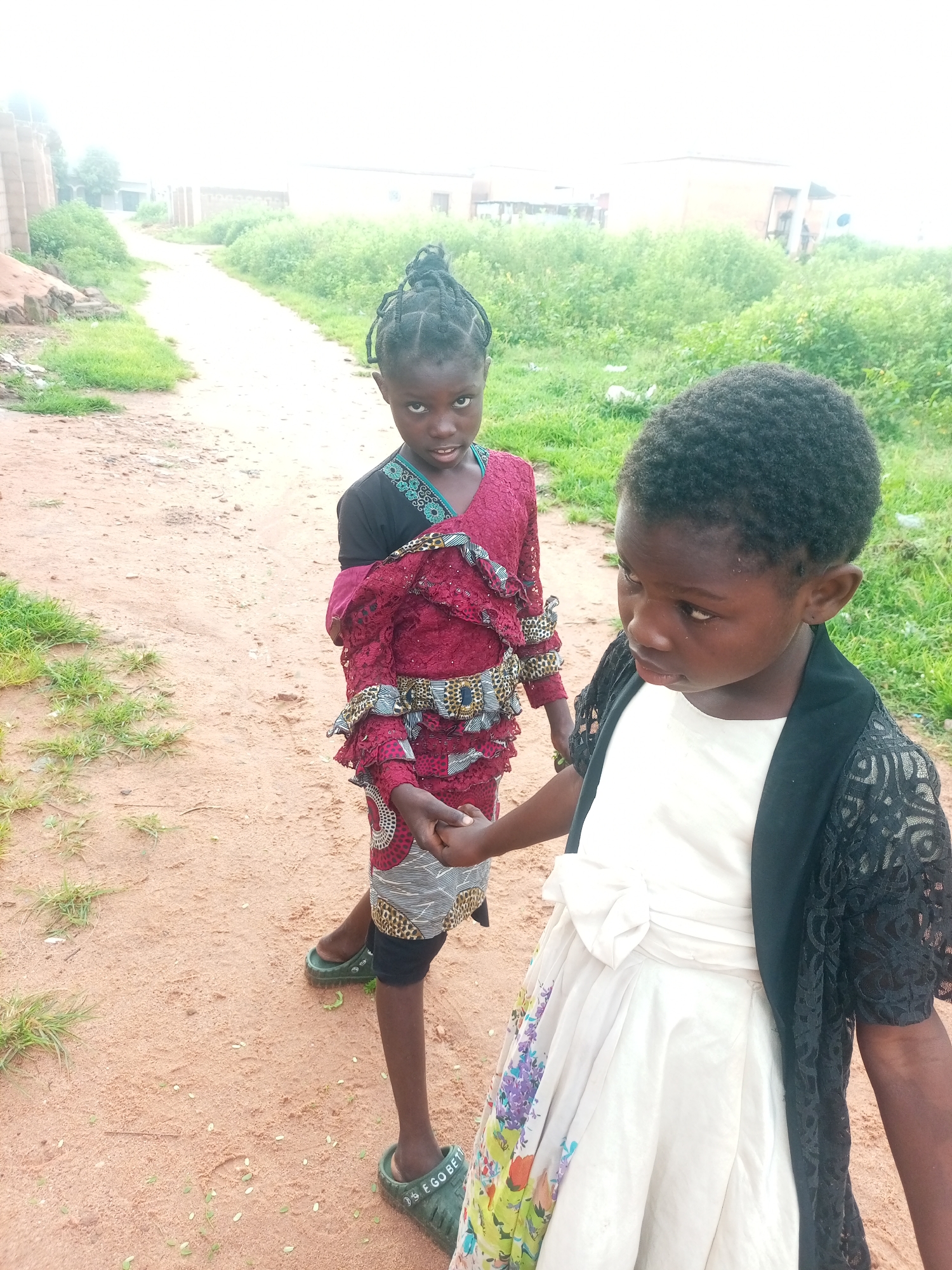 Two young girls stand on a dirt path in Tempure, Nigeria. The girl on the left, facing the camera, wears a maroon and patterned dress with black sleeves and a blue trim. Her hair is braided and styled in a bun. The girl on the right, in profile and looking down, wears a white dress with a floral print skirt and a black lace overlay. She has short, curly hair. The path is surrounded by dry grass and some scattered debris. In the background, there are low-rise buildings and more vegetation. The sky is overcast.