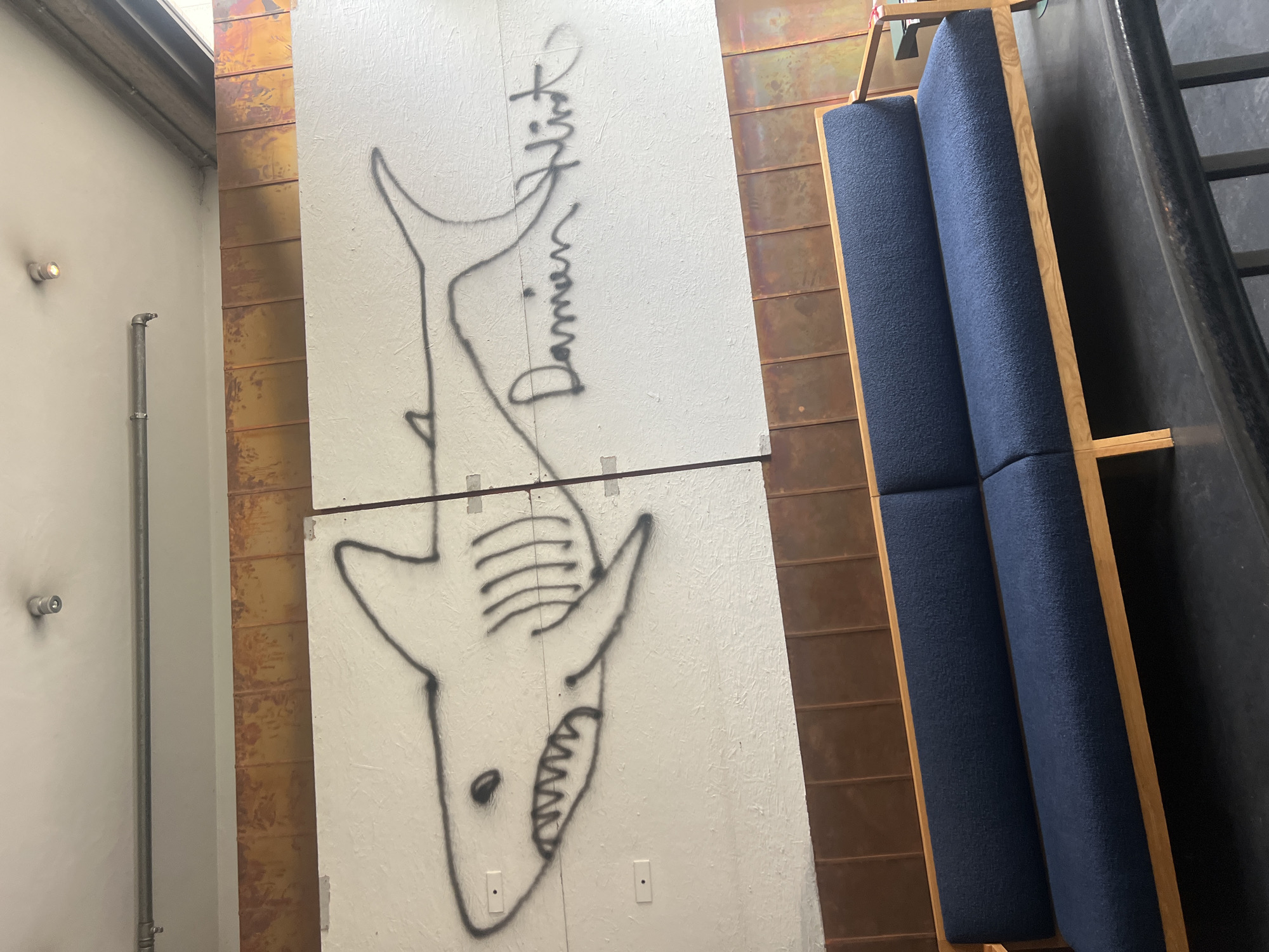 A long, dark blue sofa with a wooden frame sits in front of a white wall adorned with graffiti of a shark and the name "Damien Hirst." The shark is outlined in black spray paint, and its mouth is open, revealing jagged teeth. The name is written in a black, cursive font to the right of the shark. The wall appears to be made of two large panels. Above the white wall is a band of textured, copper-colored material. The ceiling is white and has a light fixture and a metal pipe running across it. The image was taken in Berlin, Germany.